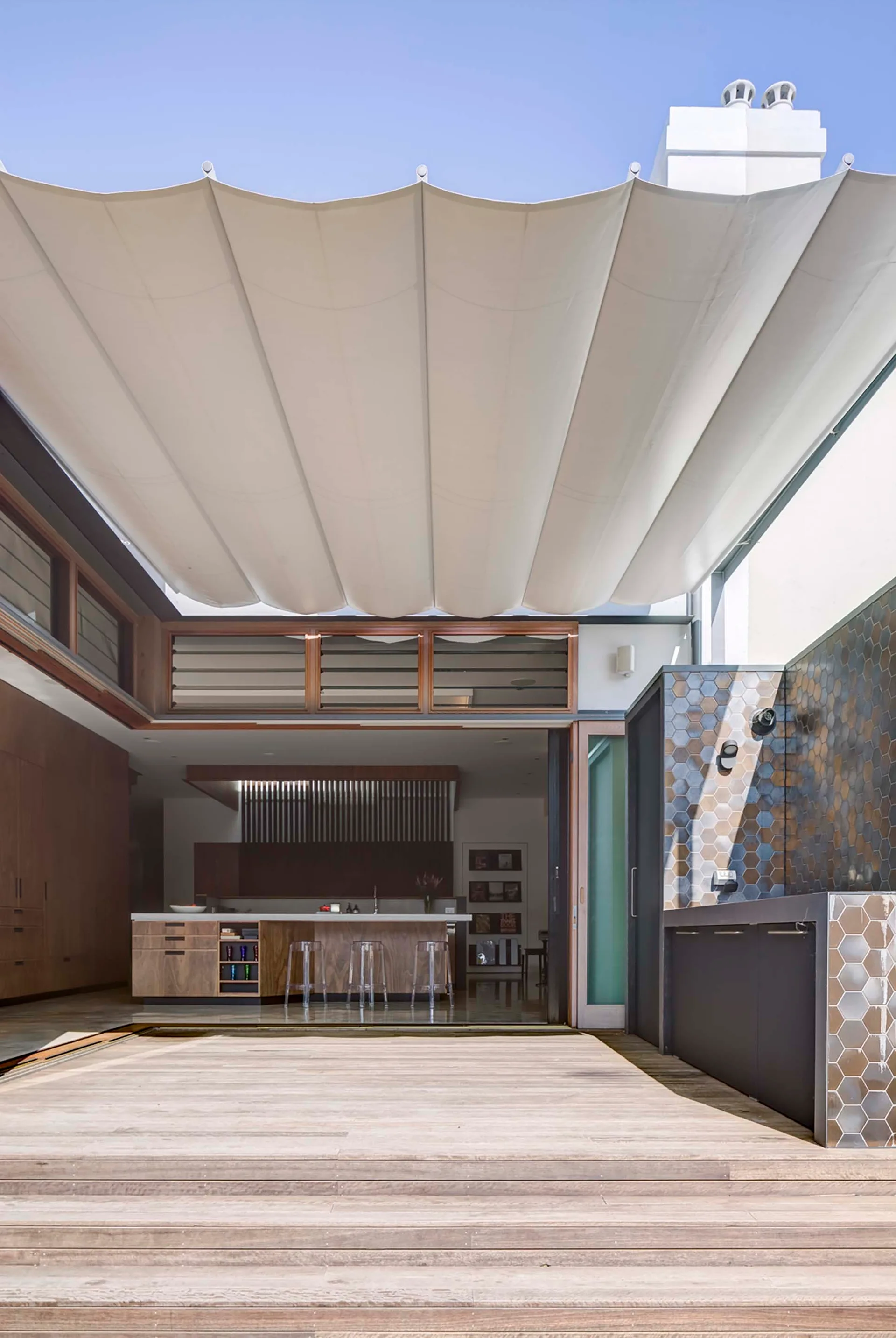 A modern wood deck patio with a white retractable fabric canopy opening into a kitchen with wood cabinetry and clear acrylic stools.