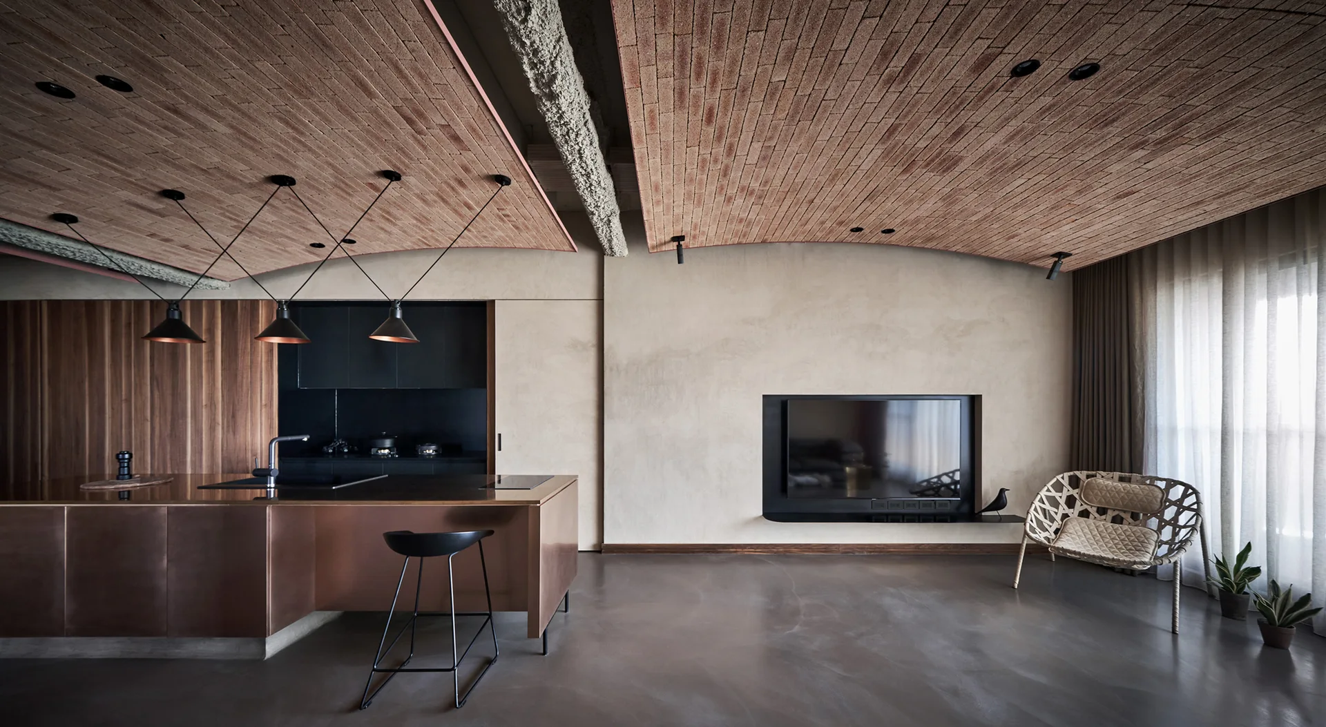 A minimalist kitchen features a metallic copper island under a vaulted brick ceiling, next to a plaster wall with a built-in television.