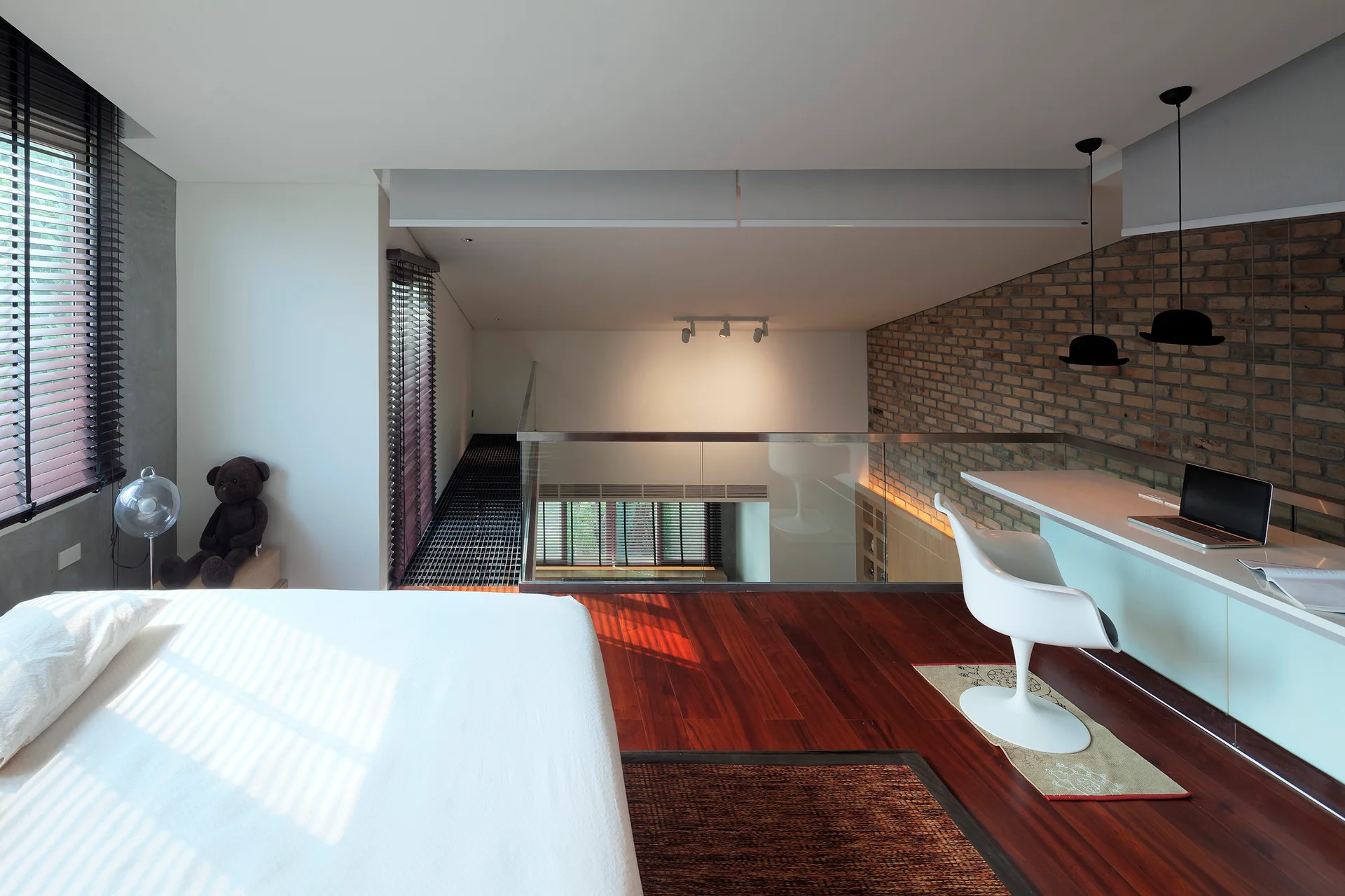 A loft bedroom featuring a bed with white bedding, a built-in desk against an exposed brick wall, and a glass railing overlooking a lower level.