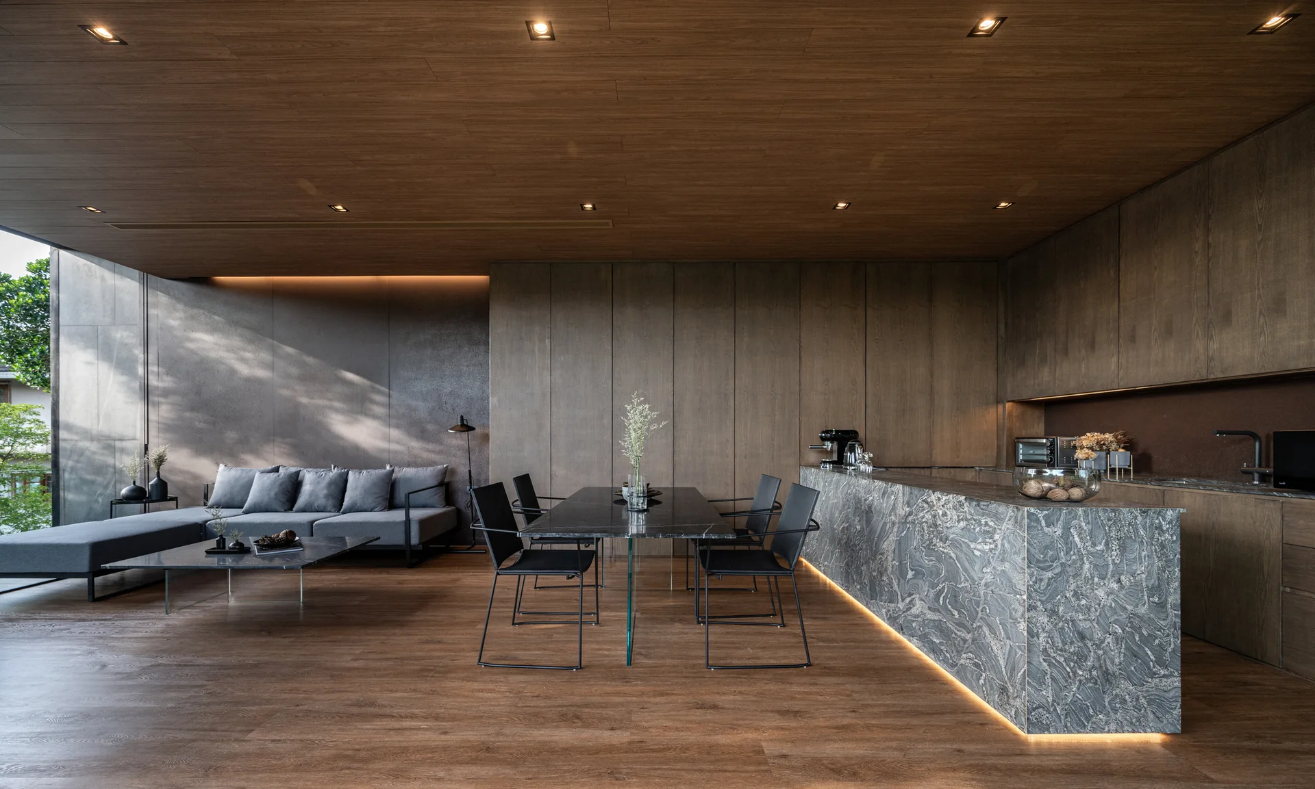 A minimalist open-plan interior featuring a gray sectional sofa, a dark dining table, and a large heavily veined gray stone kitchen island.