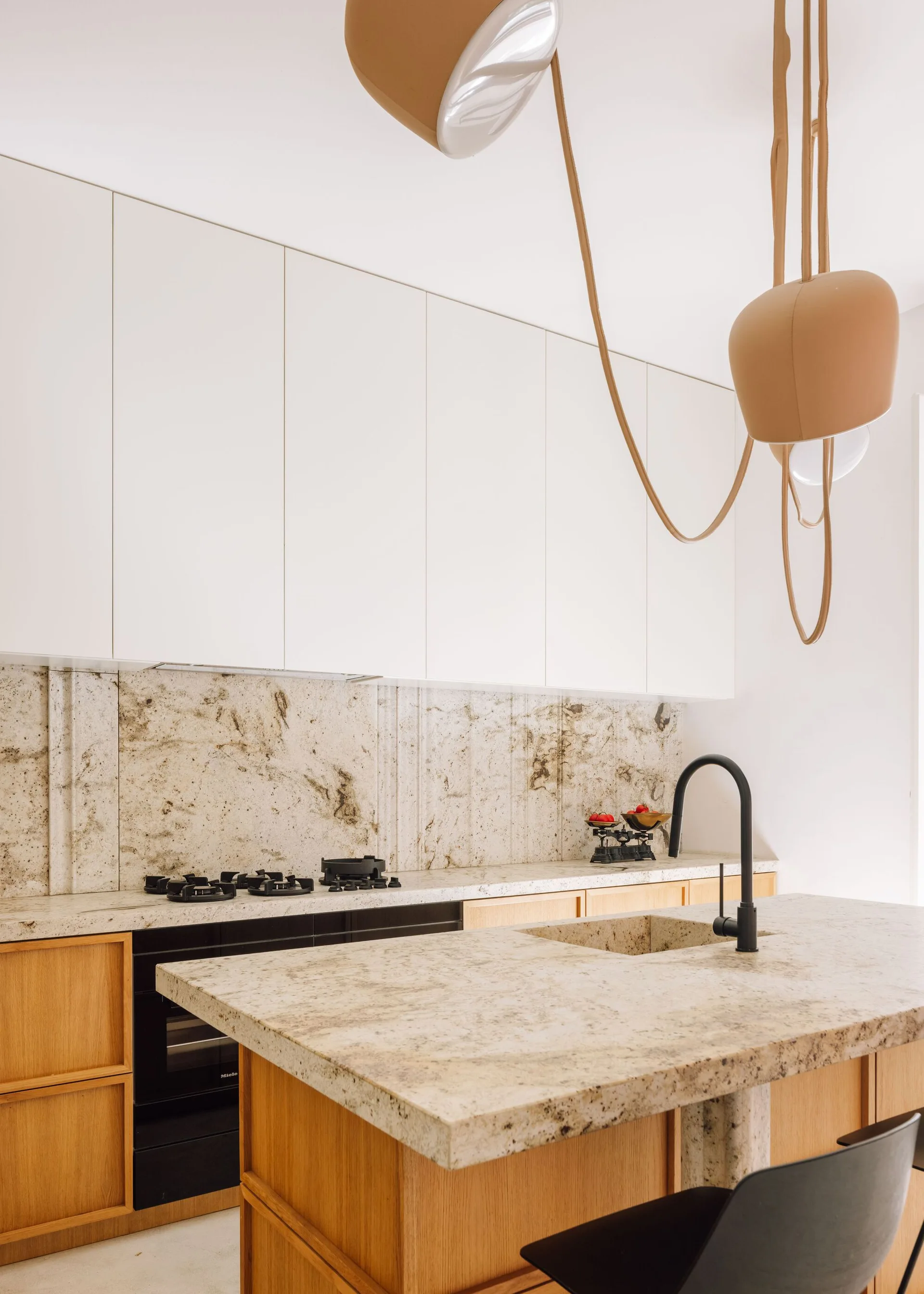 A modern kitchen featuring white upper cabinets, natural wood base cabinets, a flecked stone backsplash, and an oversized tan pendant light.