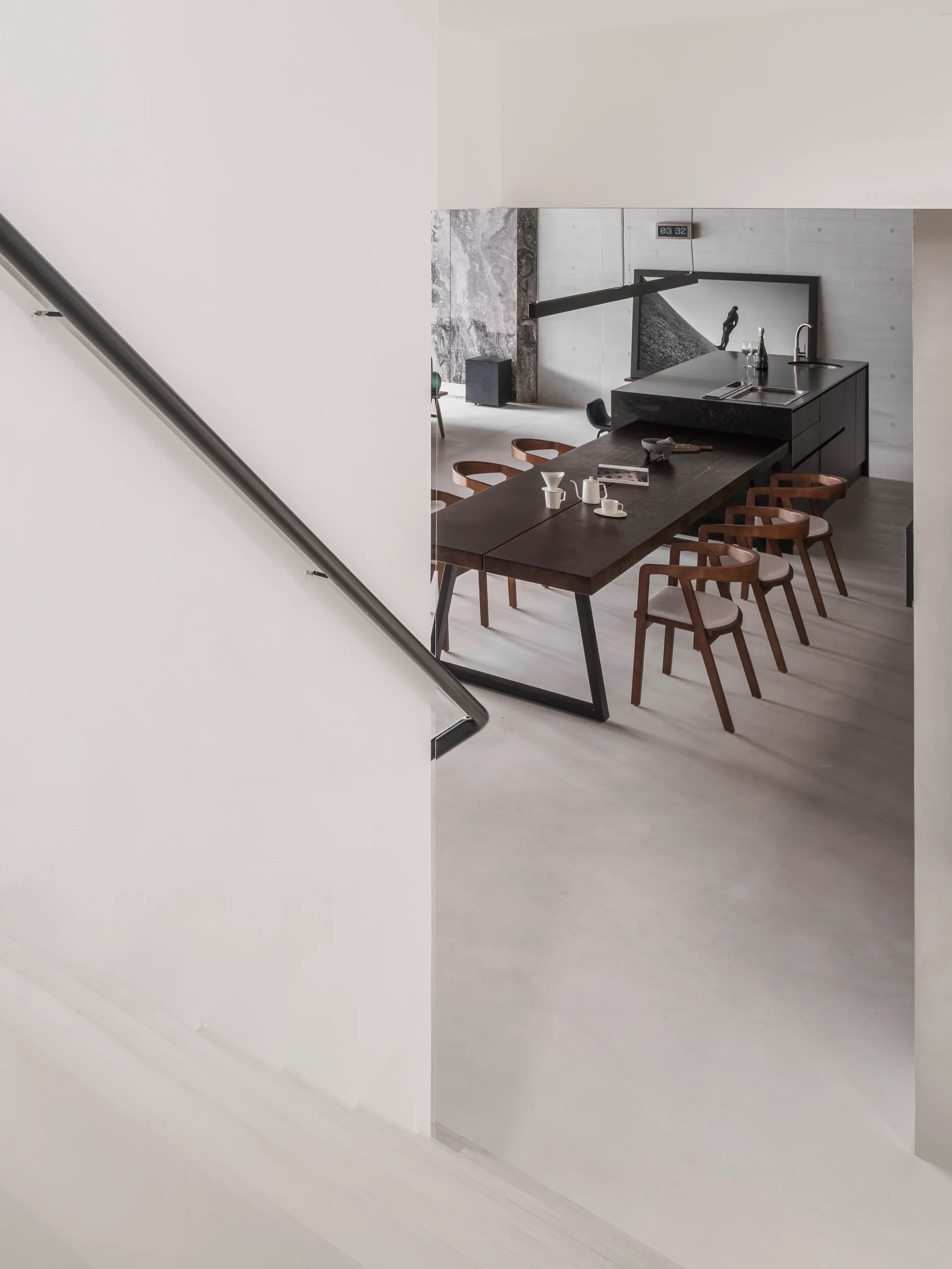 A view from a white staircase showing a dark wood dining table with chairs next to a black kitchen island against a concrete wall.