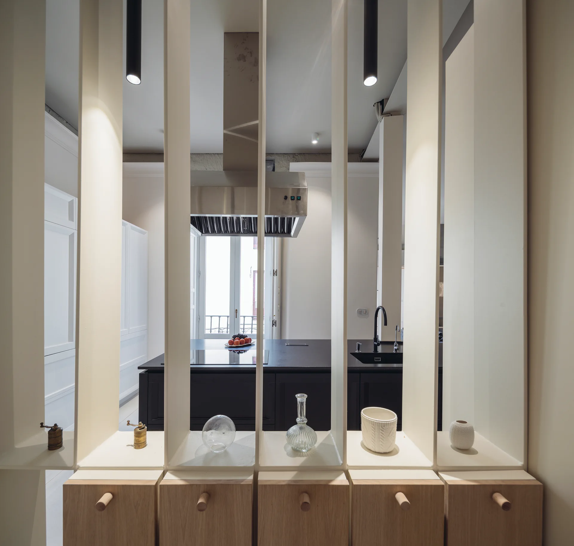 A white slatted room divider with small wooden drawers stands before a kitchen with a dark island and stainless steel vent hood.