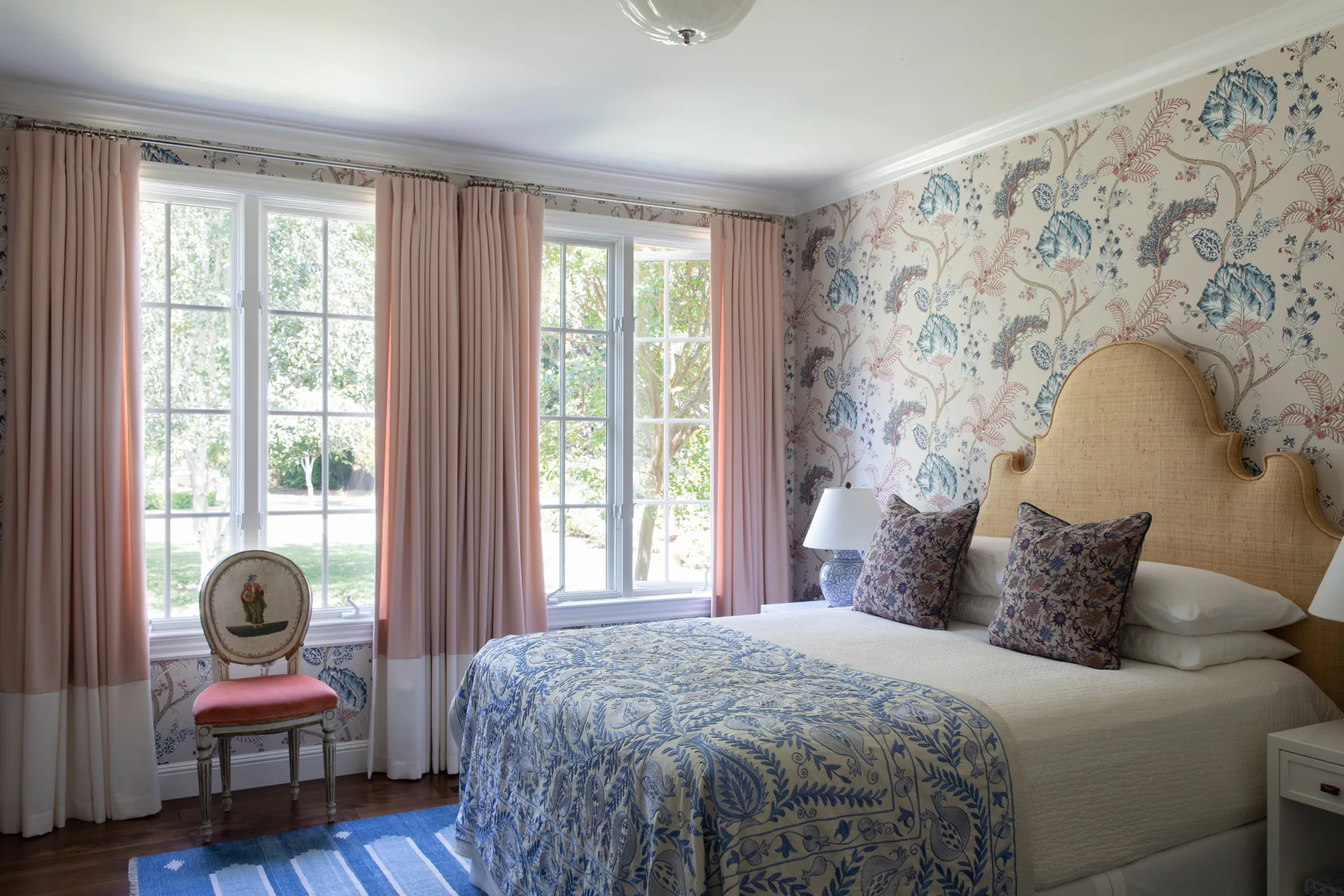 A traditional bedroom featuring botanical wallpaper, a bed with a woven headboard, pink and white curtains, and a blue patterned quilt.