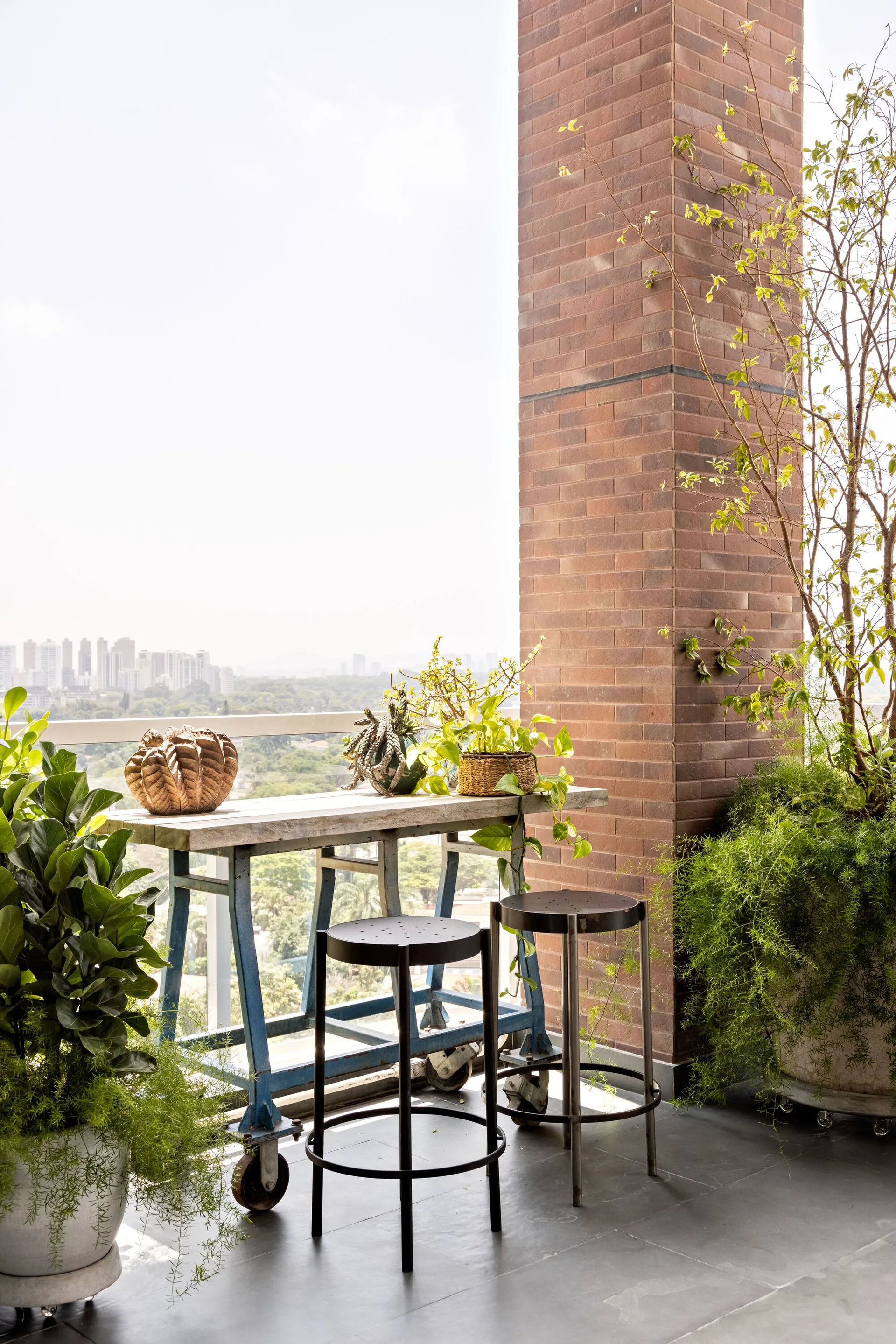 An outdoor balcony features an industrial high-top rolling table, two metal stools, potted green plants, and a tall red brick column overlooking a city view.