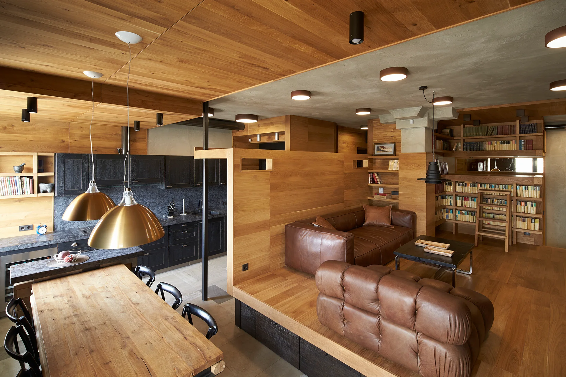 A modern open-concept room features a raised wooden living area with brown leather sofas, built-in bookshelves, and a dark kitchen.