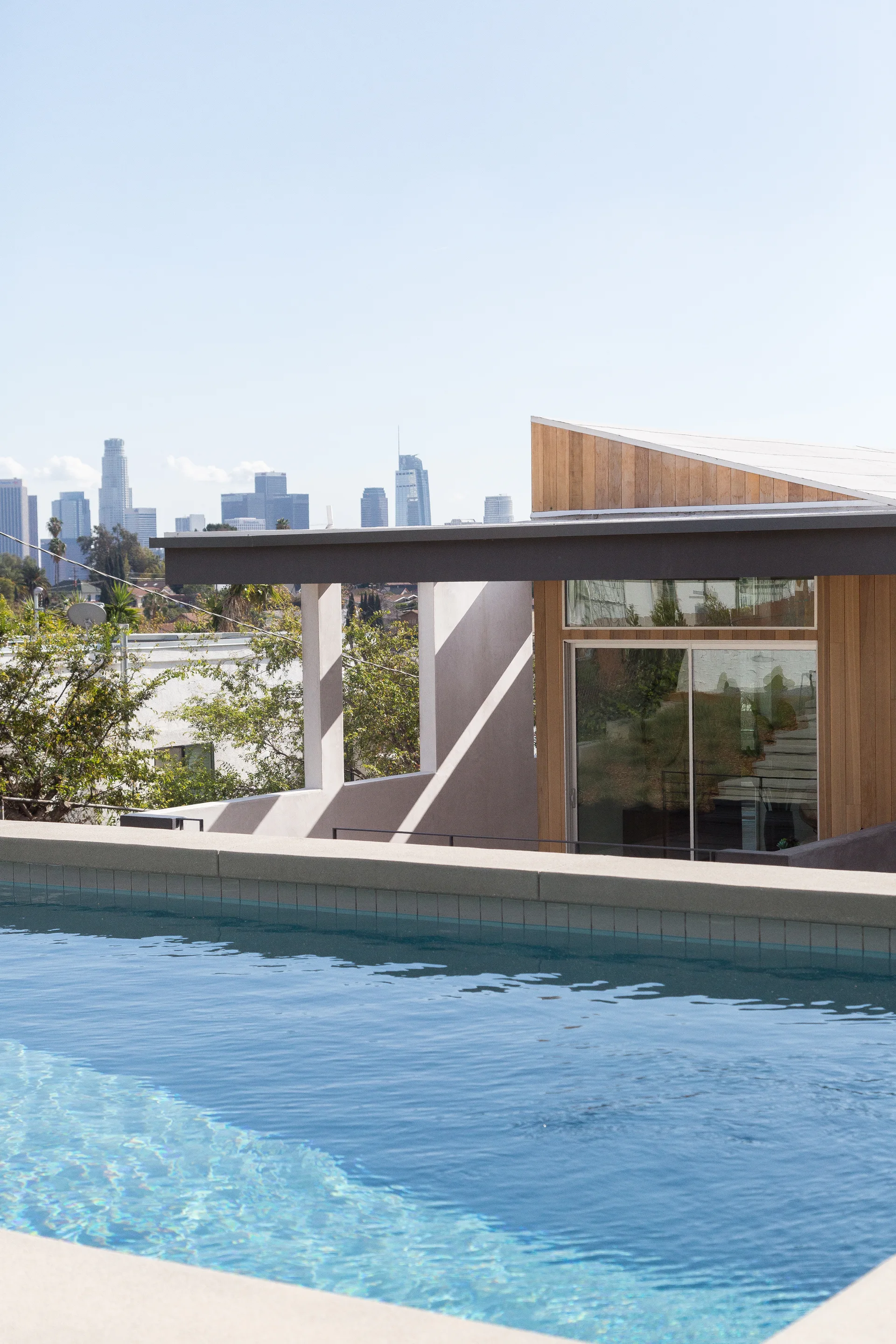 A bright blue swimming pool overlooking a modern residential building with natural wood siding and a city skyline on the horizon.