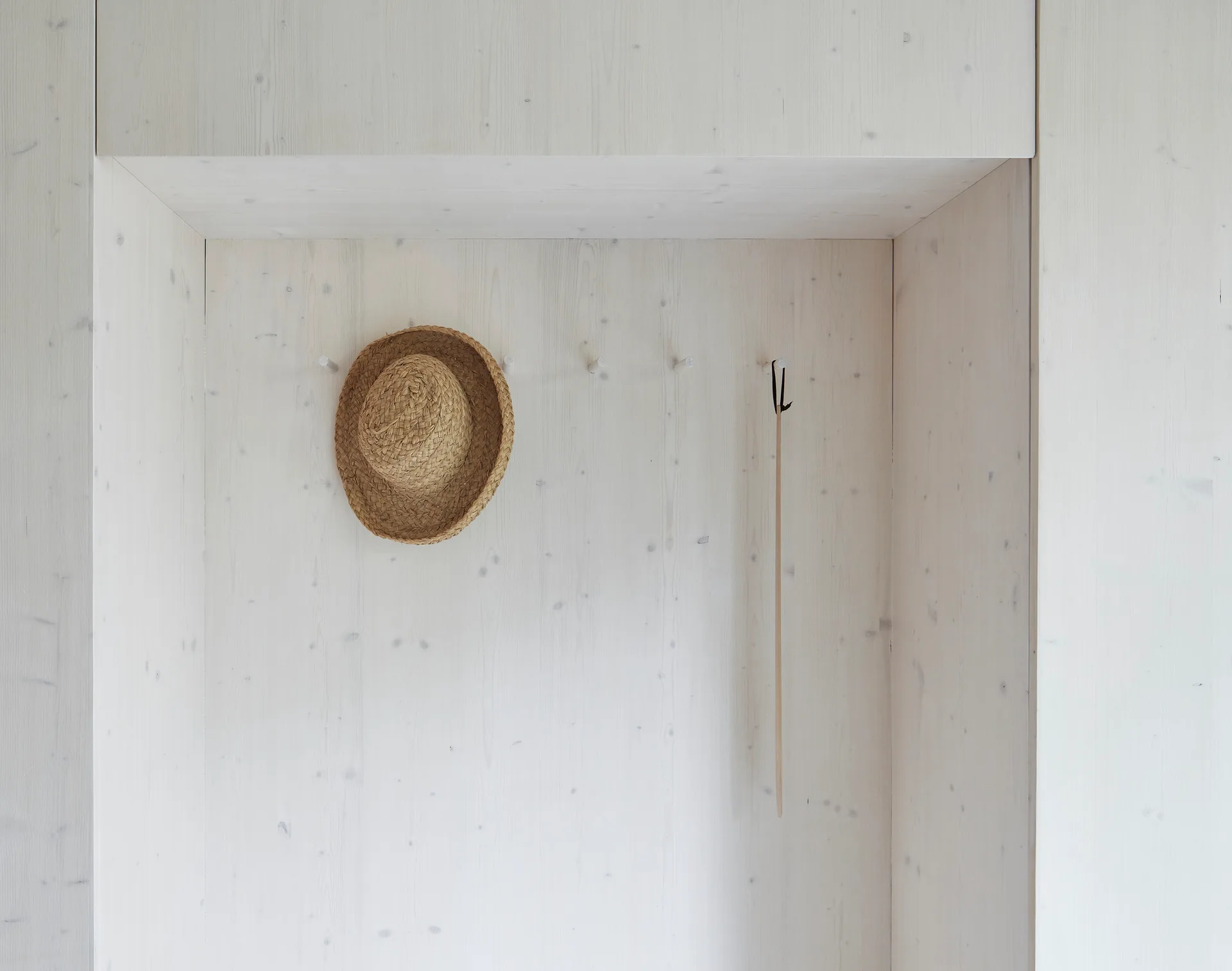 A recessed entryway alcove features whitewashed wood paneling, simple wooden wall pegs, a hanging straw hat, and a long wooden shoehorn.