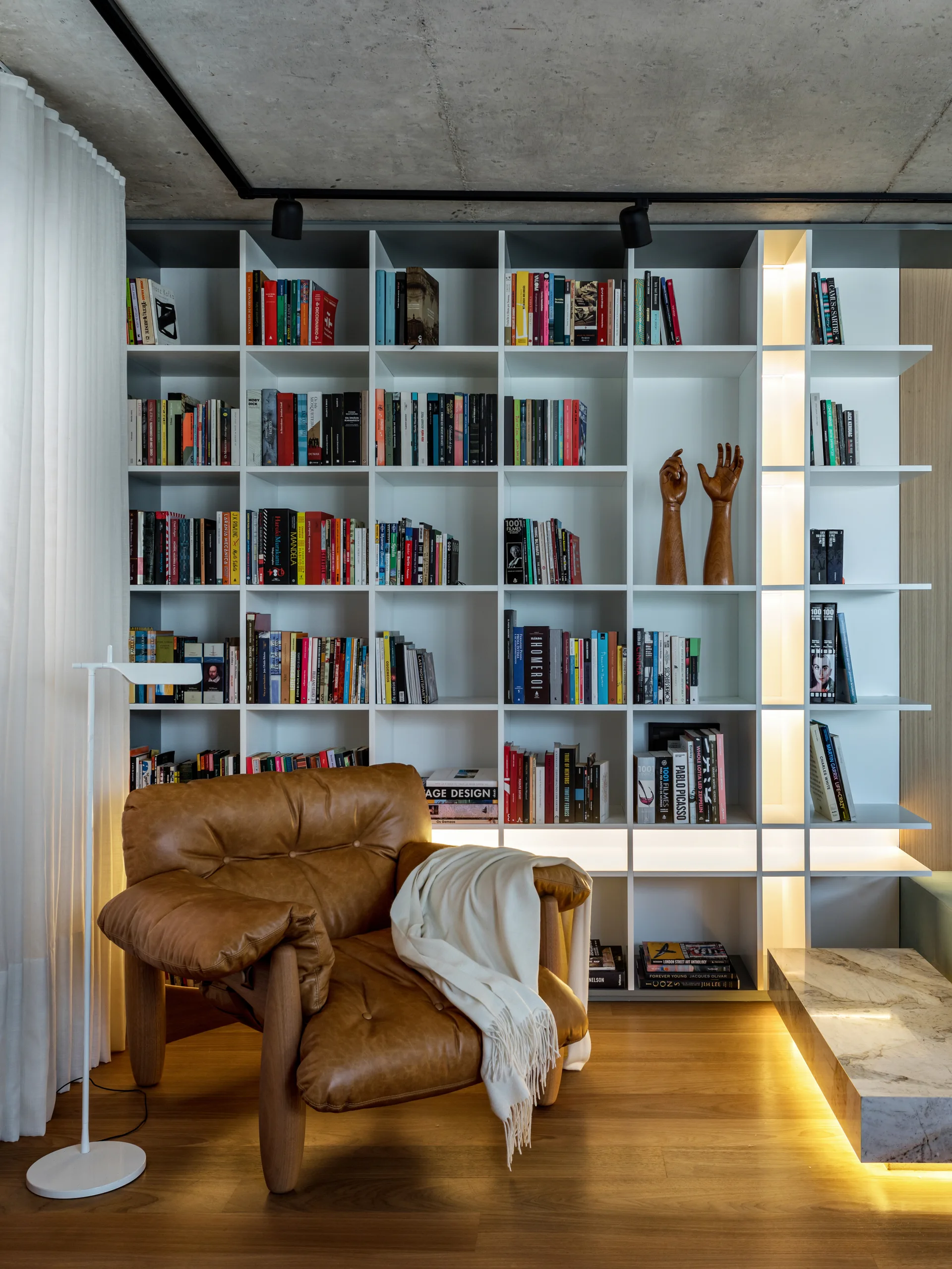 A tufted tan leather armchair with a white throw blanket sits next to a large floor-to-ceiling white bookshelf with integrated warm lighting.