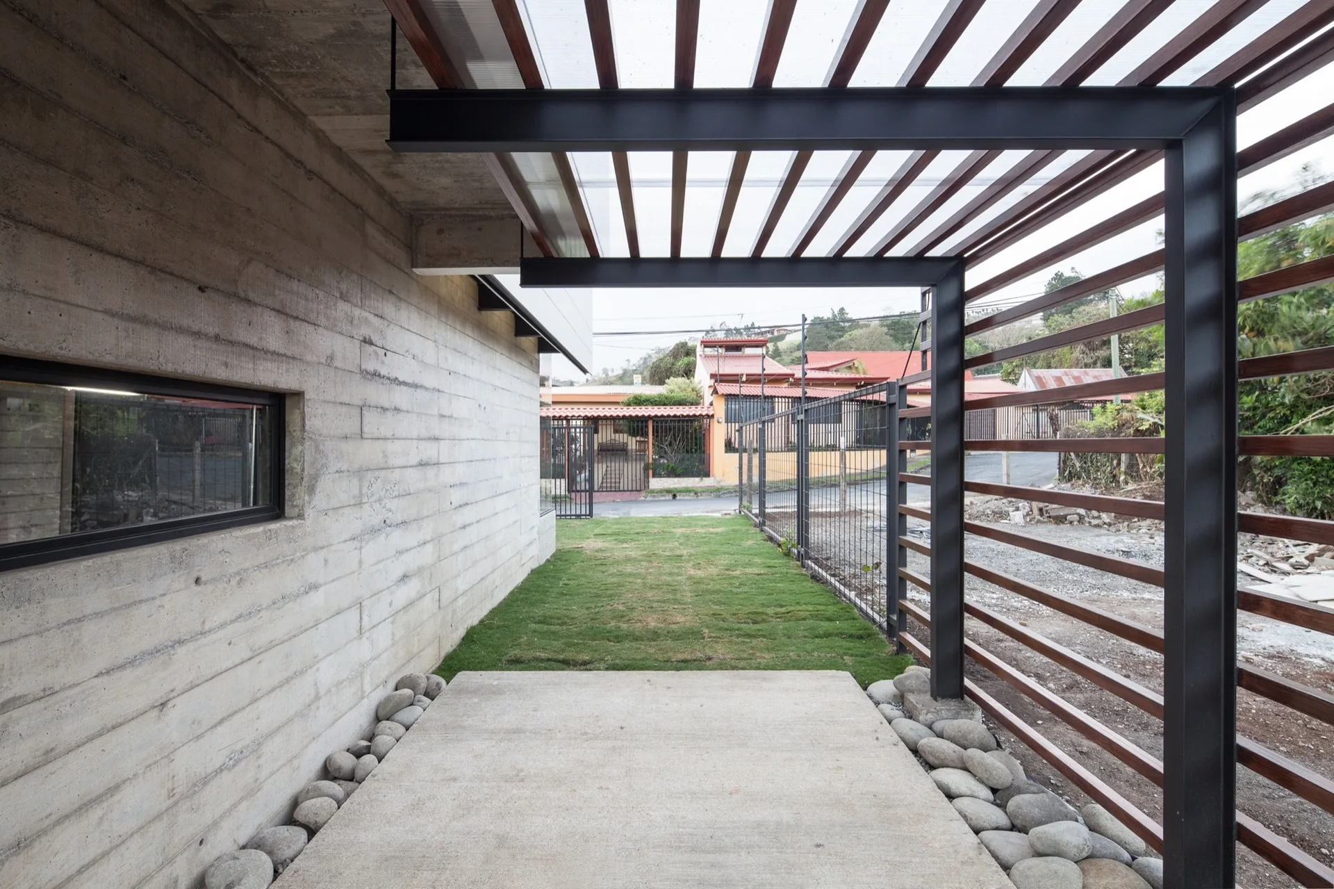 A concrete walkway bordered by river stones sits alongside a board-formed concrete wall beneath a black metal and wood slat pergola.