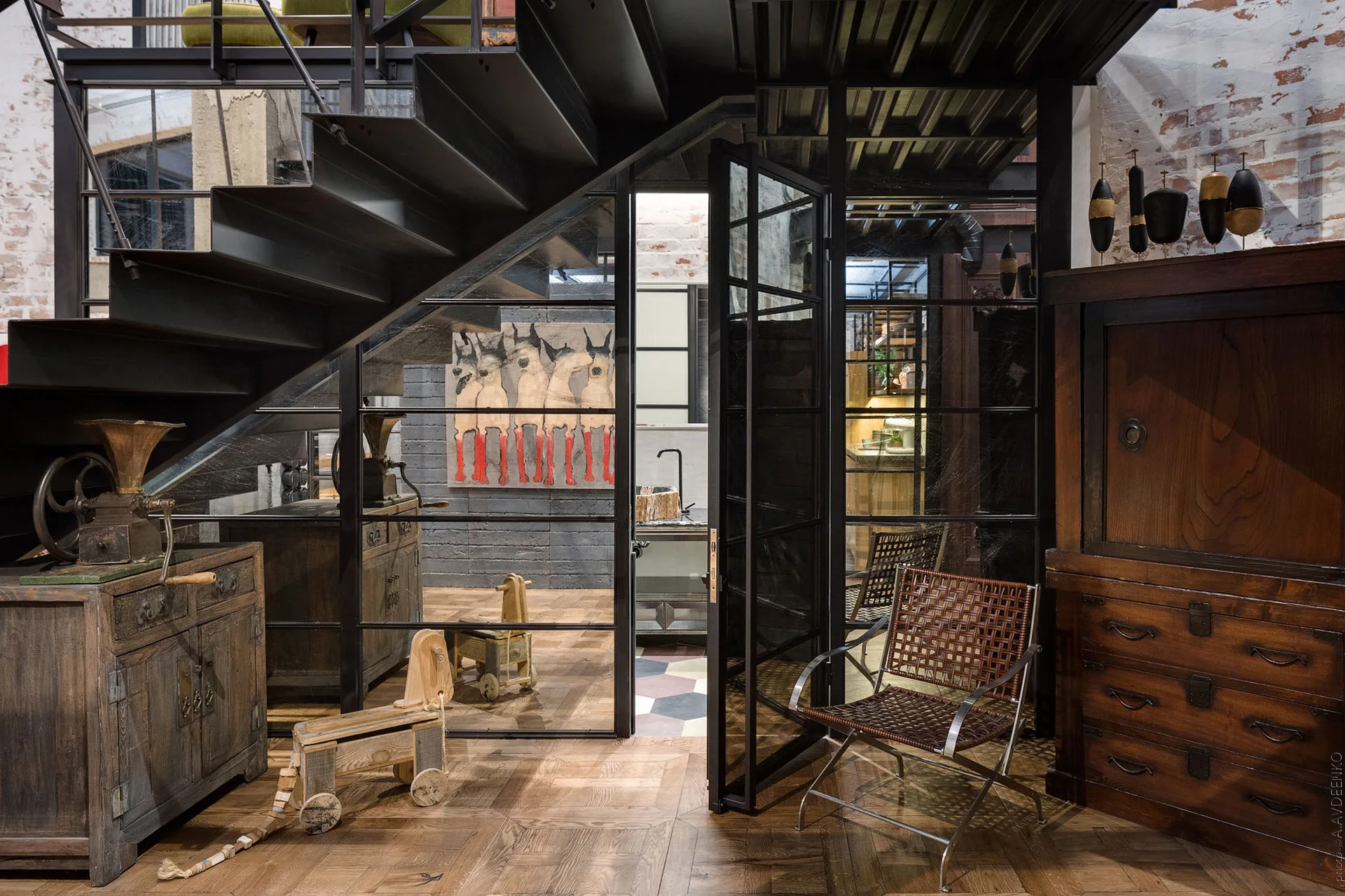 An industrial interior features a black metal staircase, a steel-framed glass partition door, and weathered wood cabinets on a wood floor.
