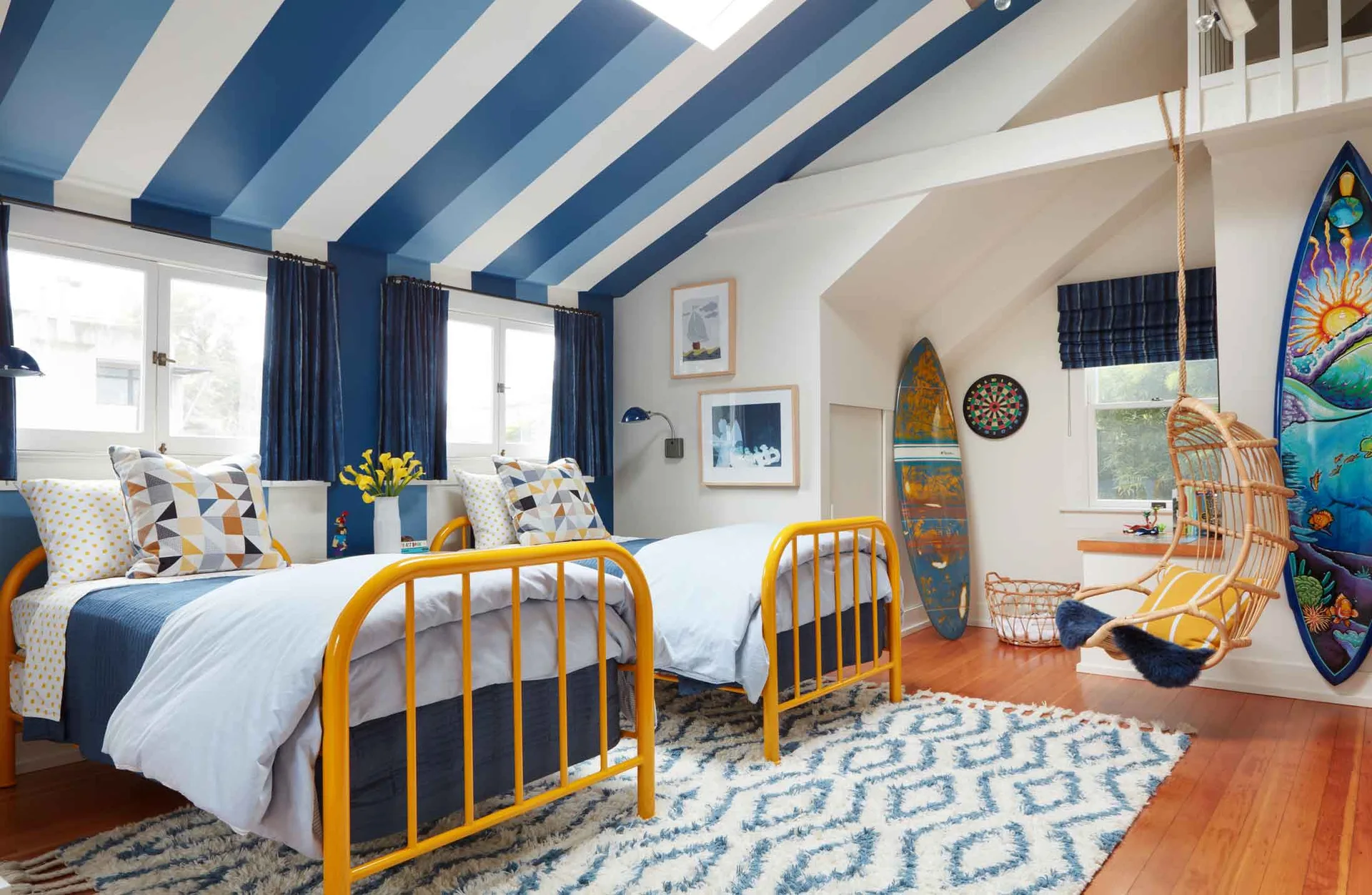 A kids' attic bedroom featuring two yellow metal twin beds, a blue and white striped ceiling, a hanging rattan chair, and decorative surfboards.