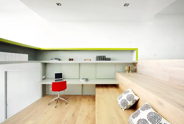 A modern minimalist home office features a grey built-in desk, a red desk chair, and stepped light wood platform seating.