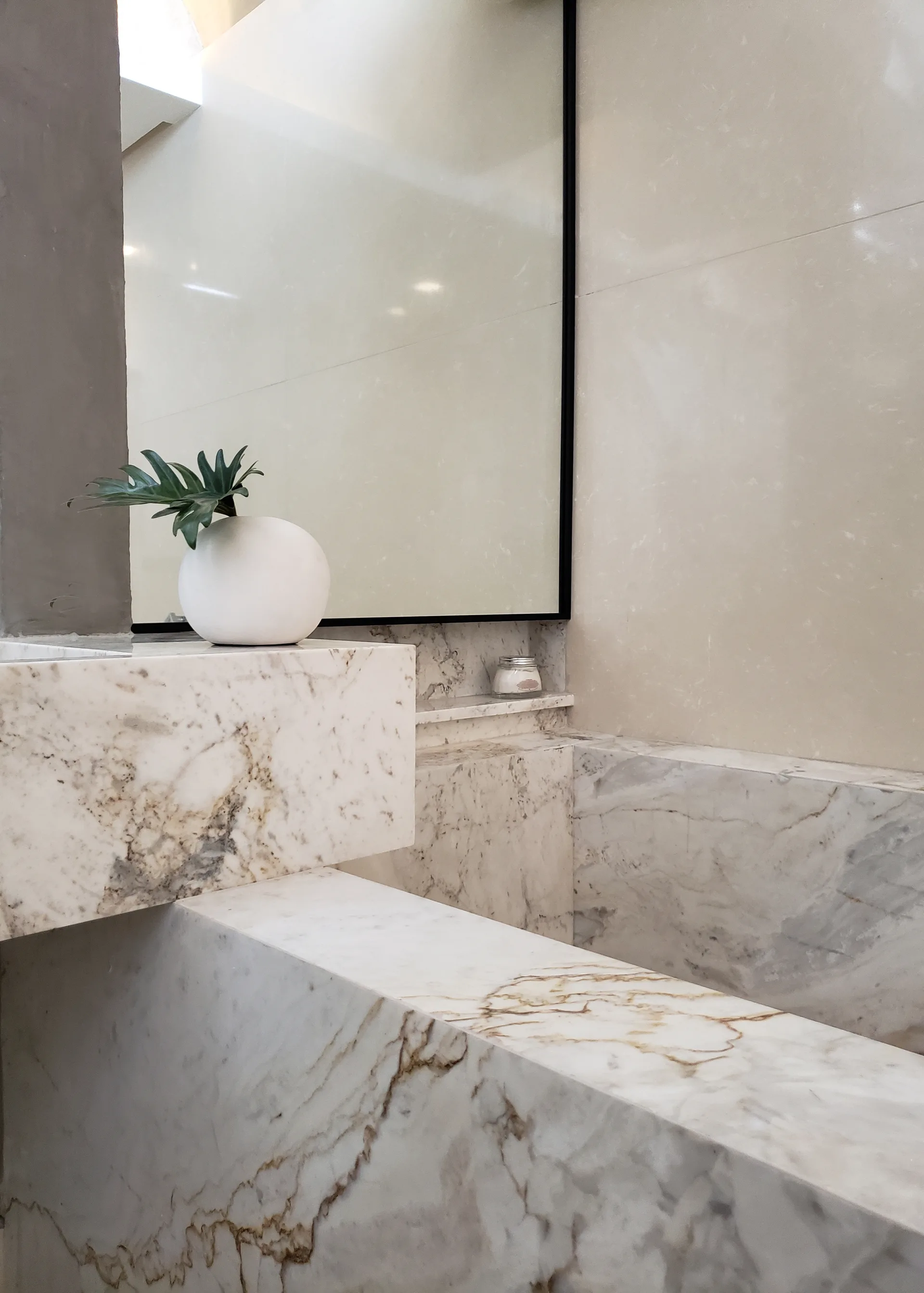 A minimalist bathroom details a cantilevered veined marble vanity, a large thin-framed mirror, and a round white vase with a single green leaf.