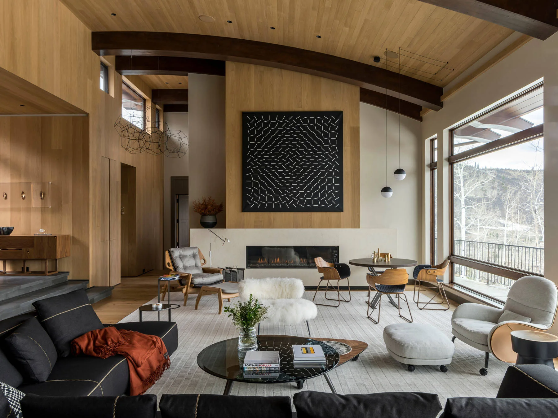 A modern living room with warm wood-paneled walls and ceiling, dark wooden beams, a linear fireplace, and large windows looking out at mountains.