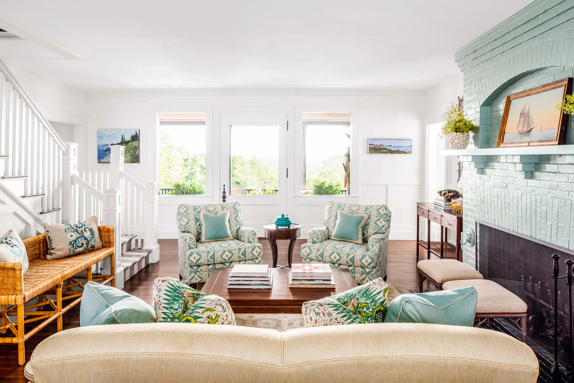 A bright living room features a mint green painted brick fireplace, two patterned teal armchairs, a central wood coffee table, and a white staircase.