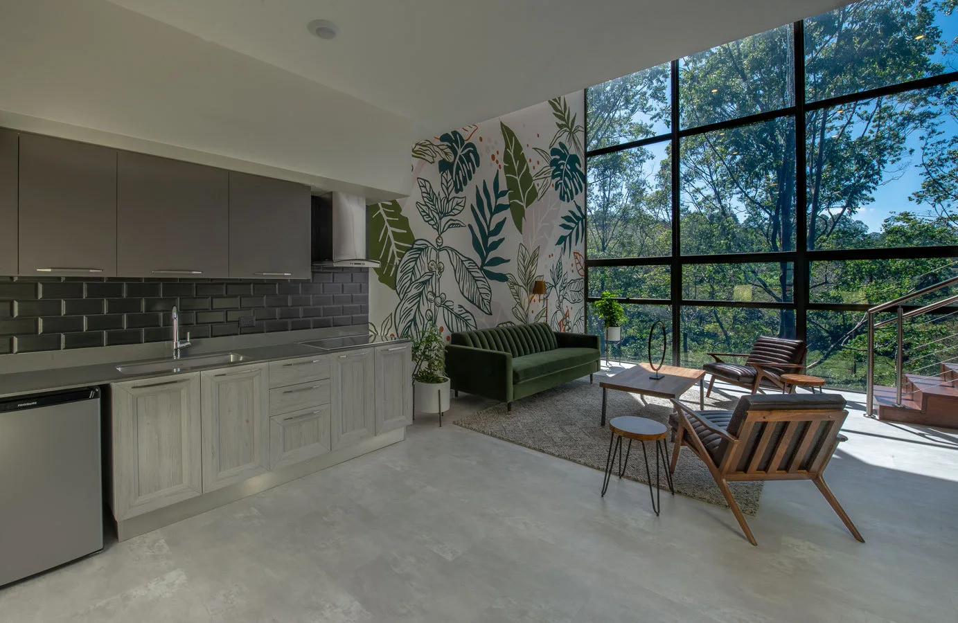 An open-concept living room and kitchen featuring a green velvet sofa, a botanical mural, and a large window facing a forest.