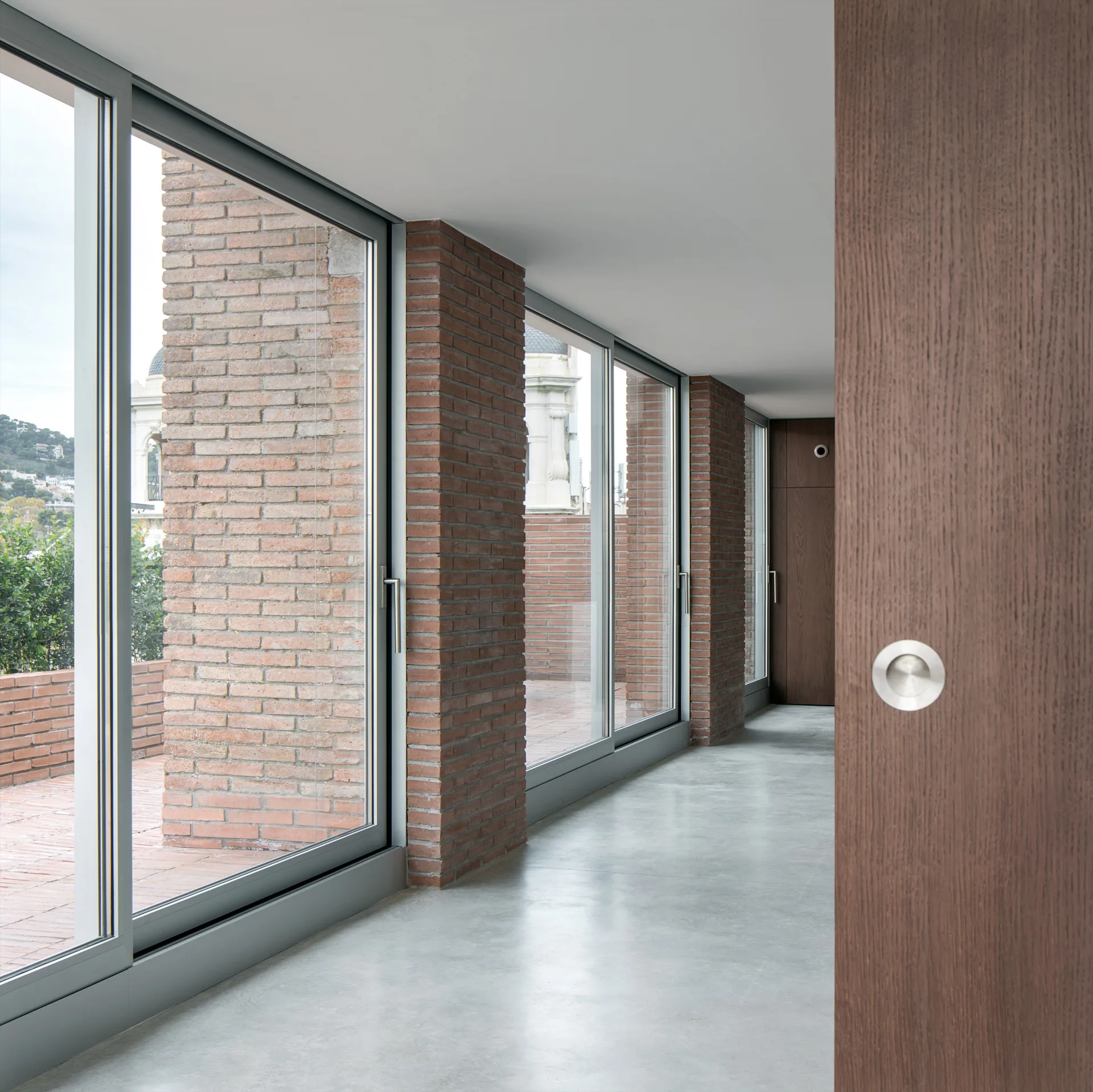 A modern hallway featuring smooth light gray concrete flooring, floor-to-ceiling glass doors separated by exposed brick columns, and a wood door.