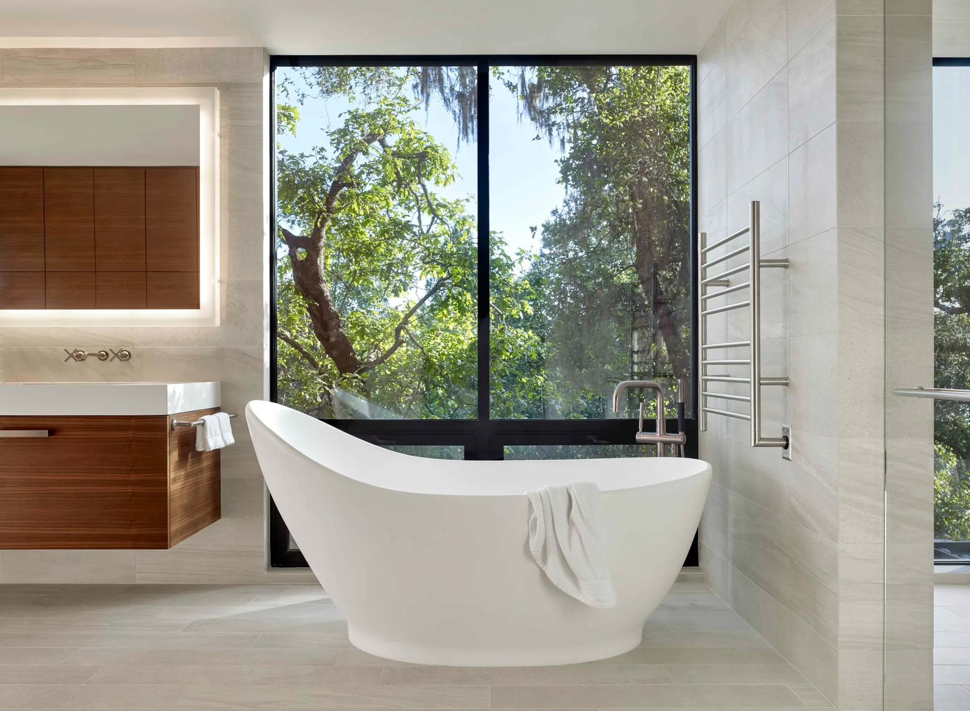A modern bathroom features a white curved freestanding bathtub in front of a large black-framed window overlooking green trees.