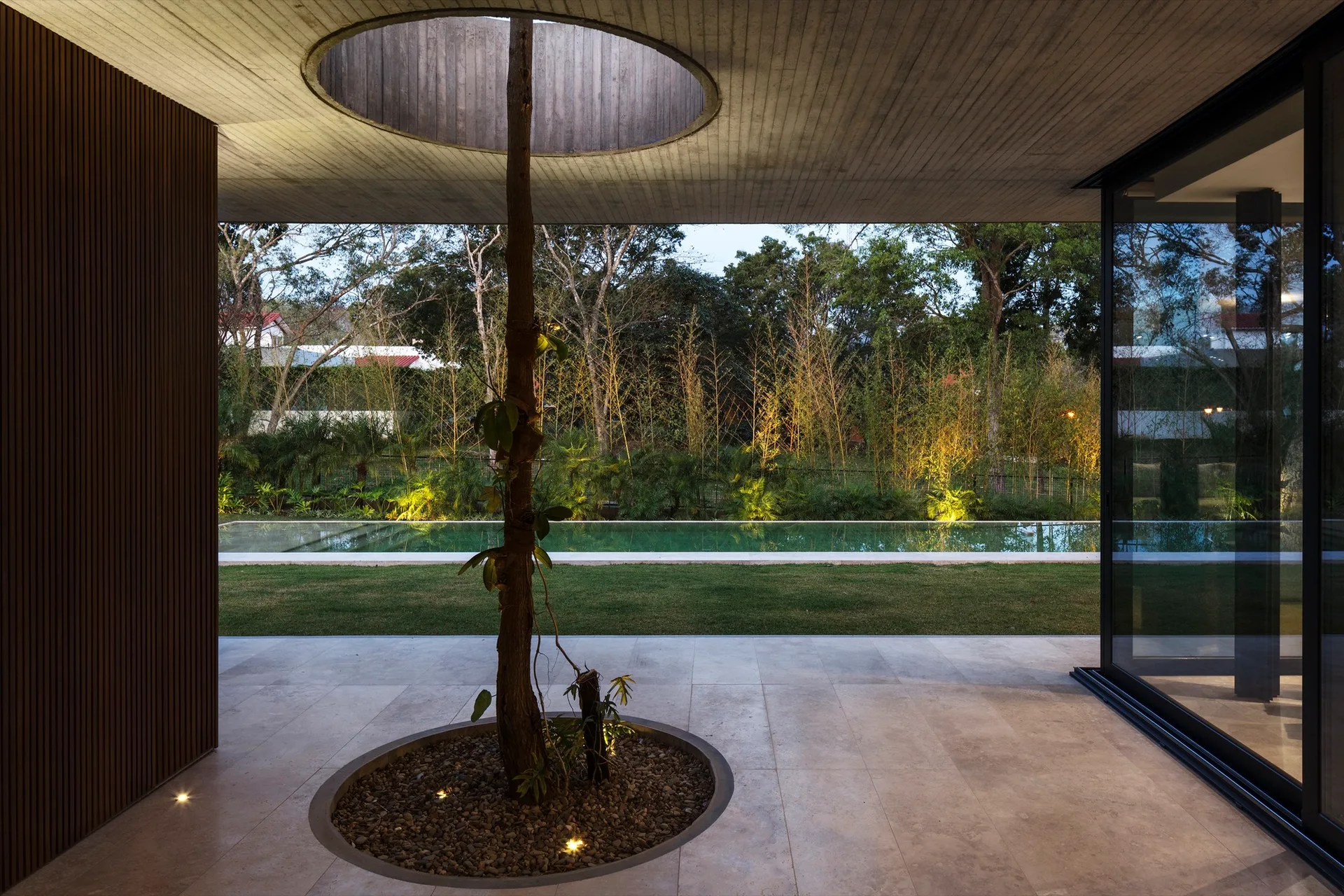 A modern covered patio features a tree growing through a circular ceiling cutout from a gravel planter, with a swimming pool visible in the background.