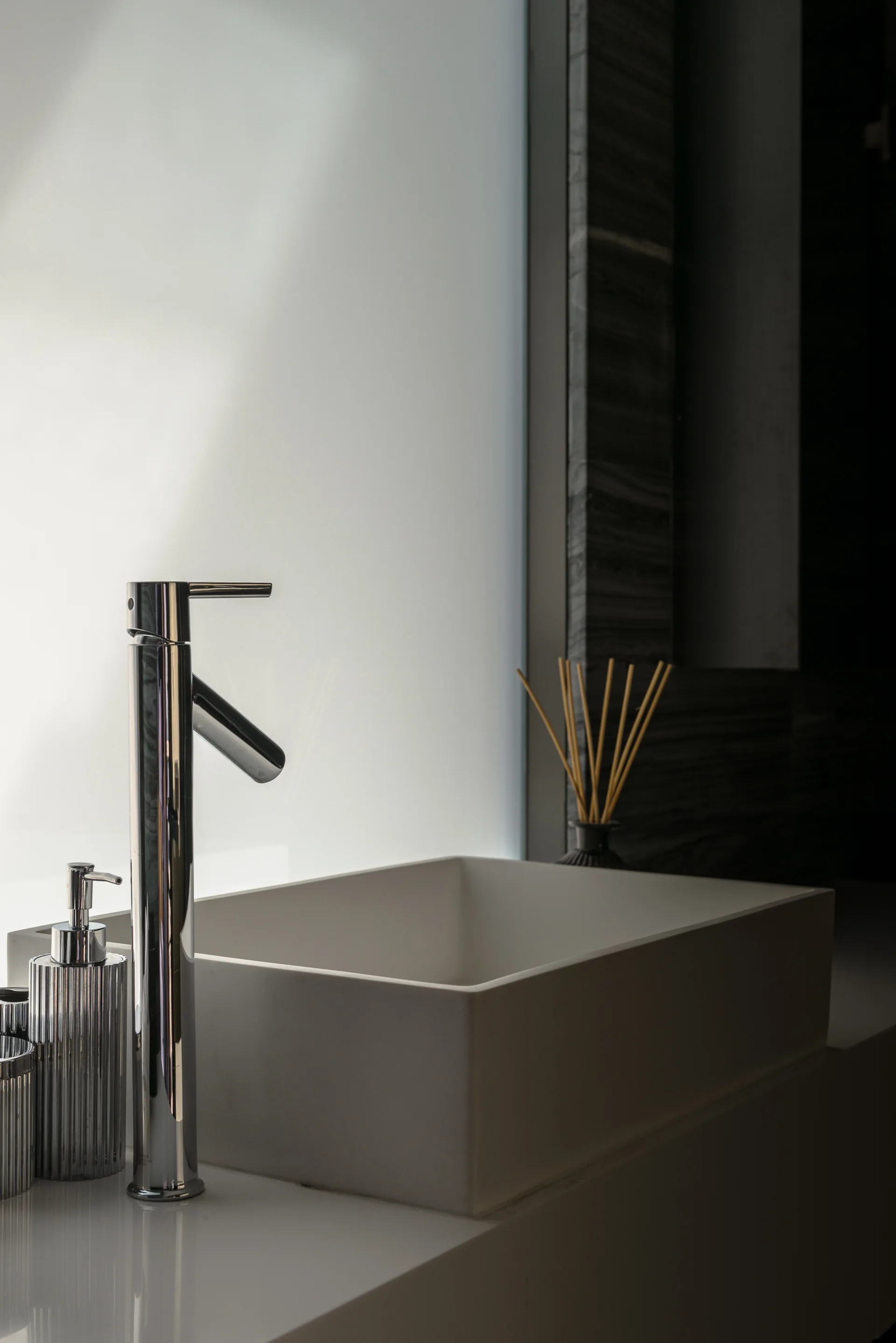 A close-up of a rectangular white vessel sink and a tall chrome faucet on a white vanity counter in front of a frosted glass window.