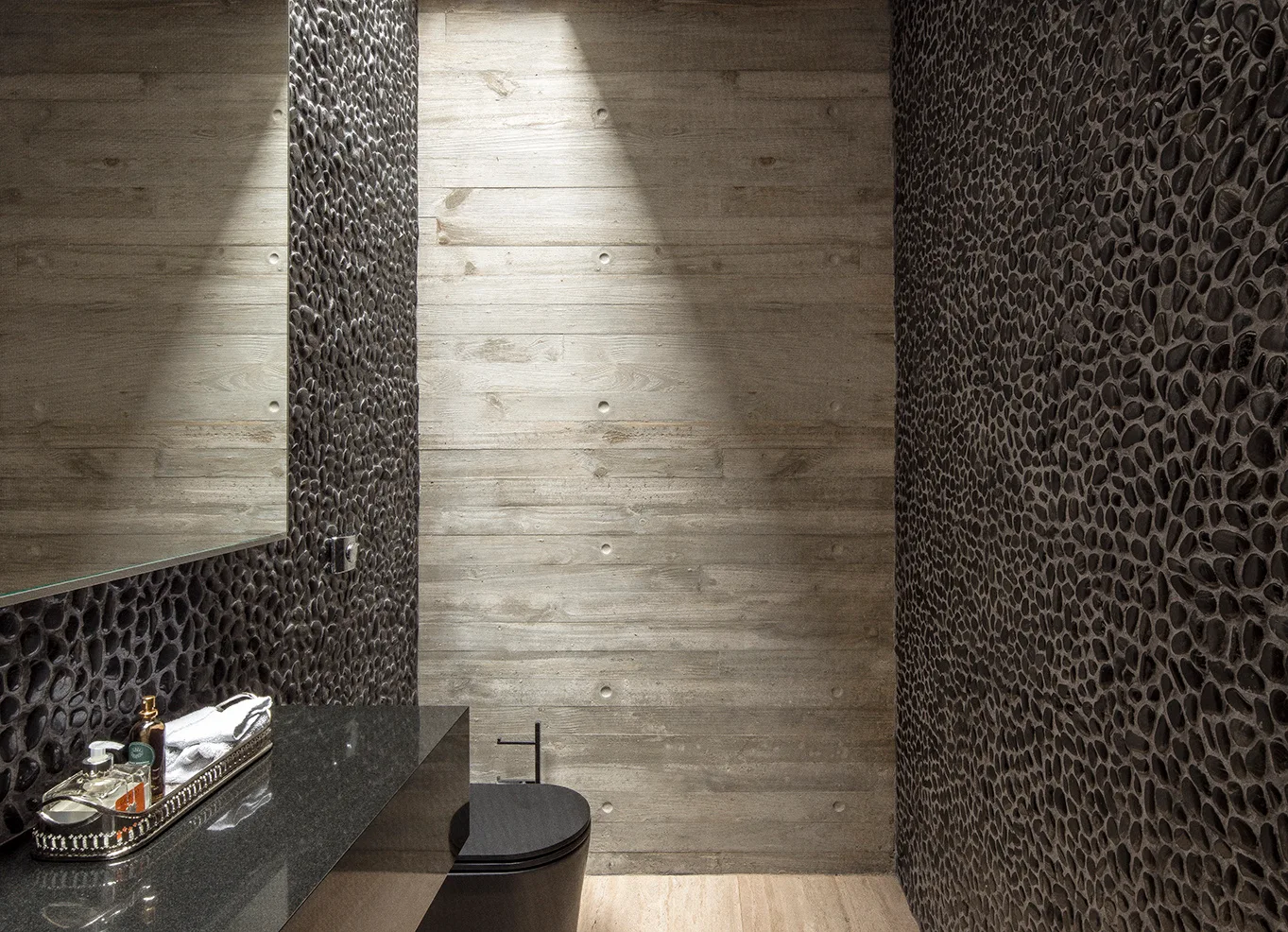 Modern powder room showing black pebble walls, a board-formed concrete back wall under a spotlight, a black toilet, and a polished dark vanity.