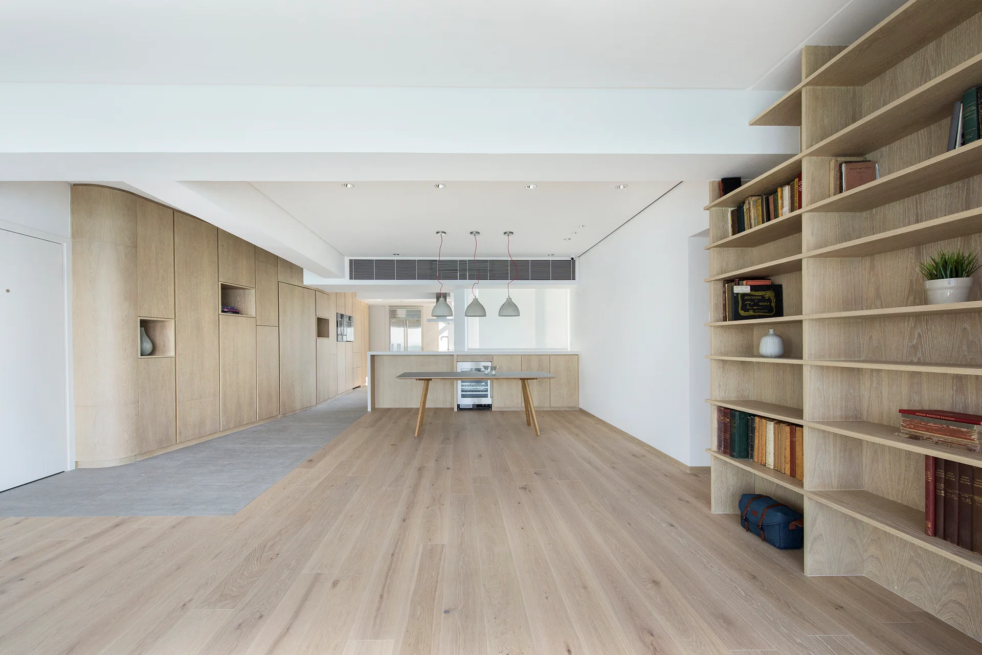 A minimalist open-plan living room features light wood floor-to-ceiling cabinetry, a curved corner wall, open bookshelves, and light-colored wood floors.