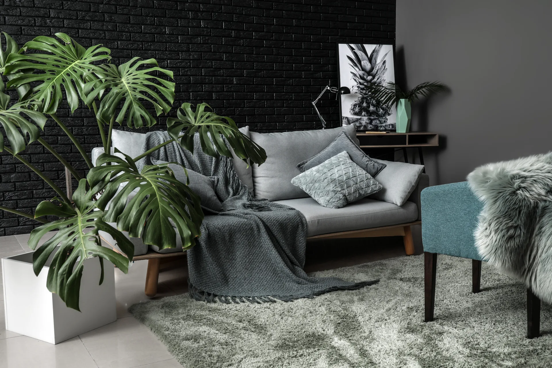 A light grey sofa sits in front of an exposed black brick wall, next to a large potted monstera plant and a teal armchair.