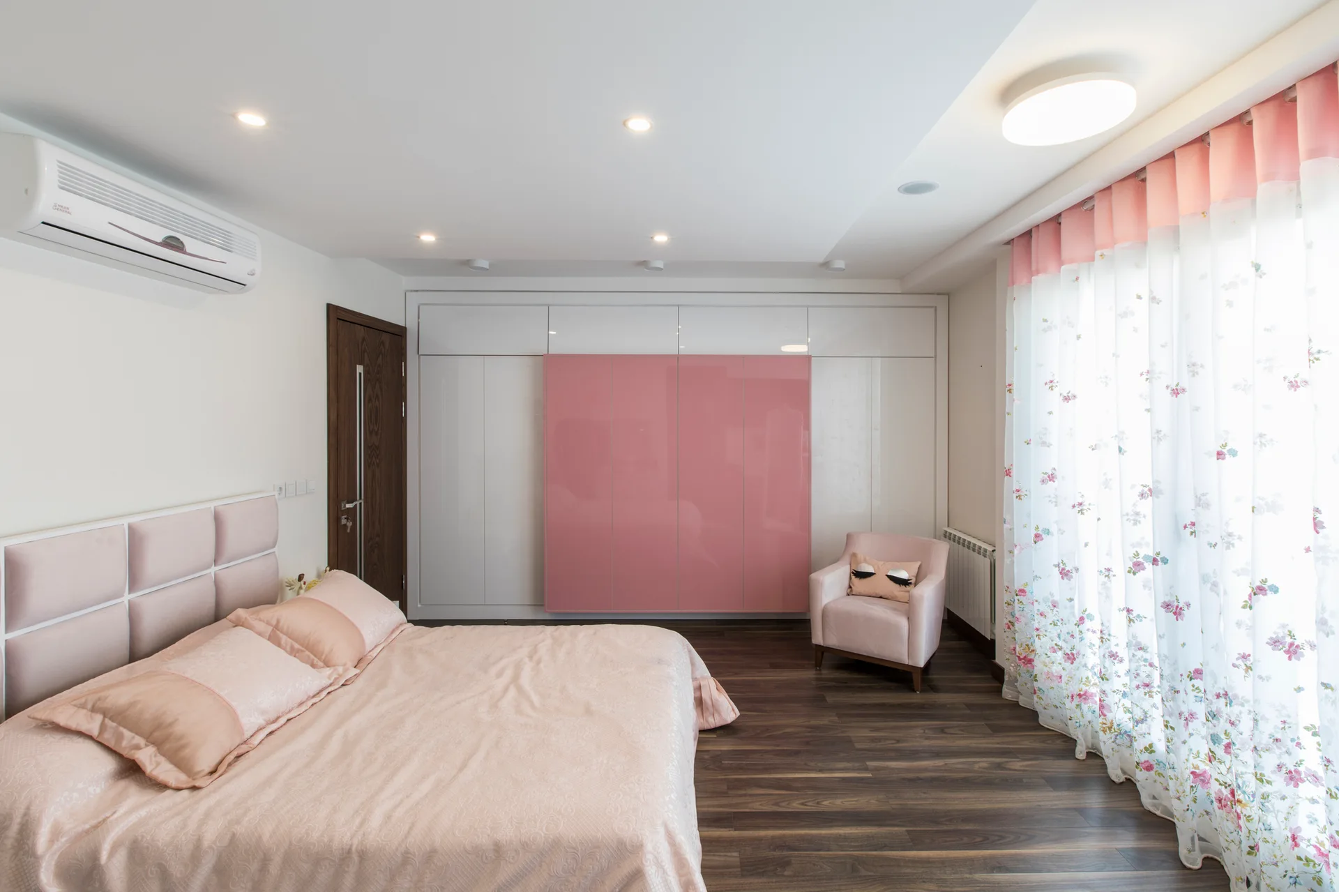 A modern white bedroom features a pink bed, a dark wood floor, sheer floral curtains, and a large built-in wardrobe with a glossy pink center panel.