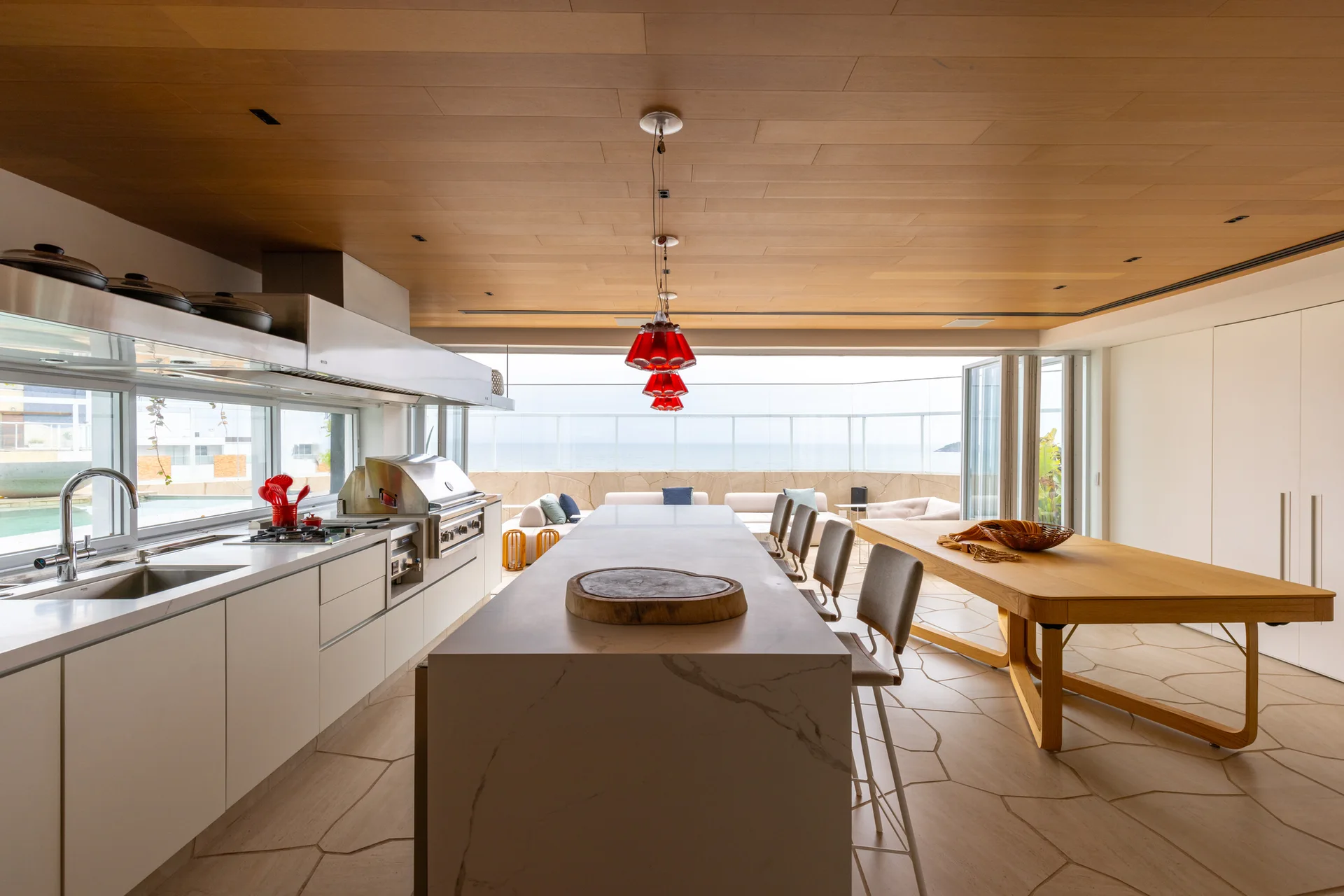 A modern kitchen featuring a marble island, a long wood dining table, a wood plank ceiling, and red pendant lights opening to an outdoor terrace.