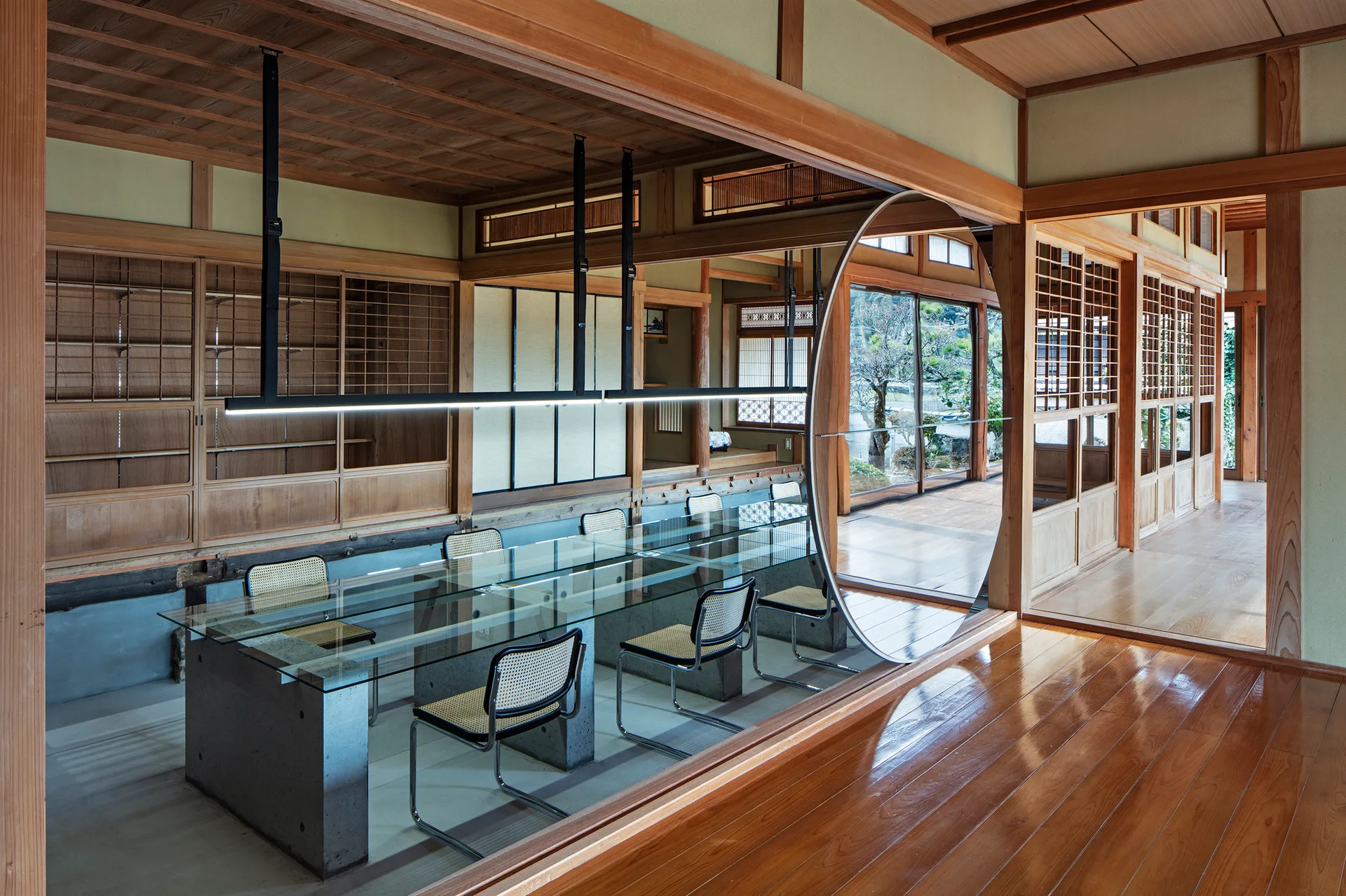 A modern glass-topped dining table with concrete bases and cane chairs is set within a traditional Japanese room featuring wooden paneling and shoji screens.