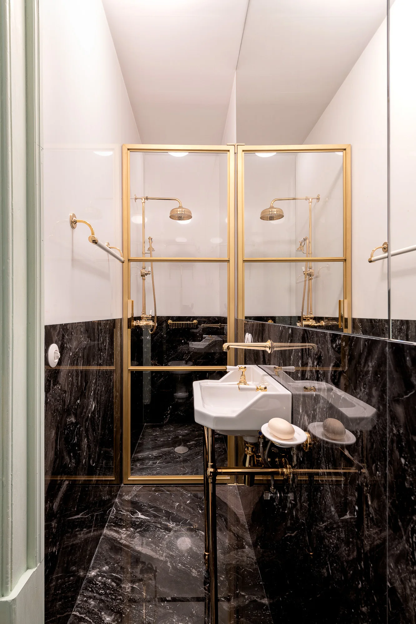 A compact bathroom featuring black veined marble flooring and wainscoting, a pedestal sink on a brass stand, and a glass-enclosed shower.