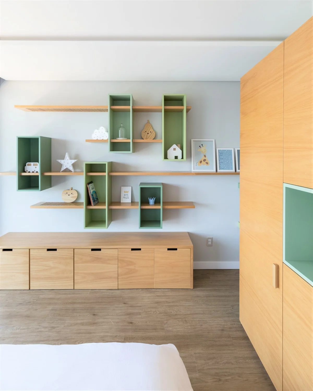 A children's room features custom light wood storage cabinets, a tall wardrobe, and modular wall-mounted shelves with sage green accents.