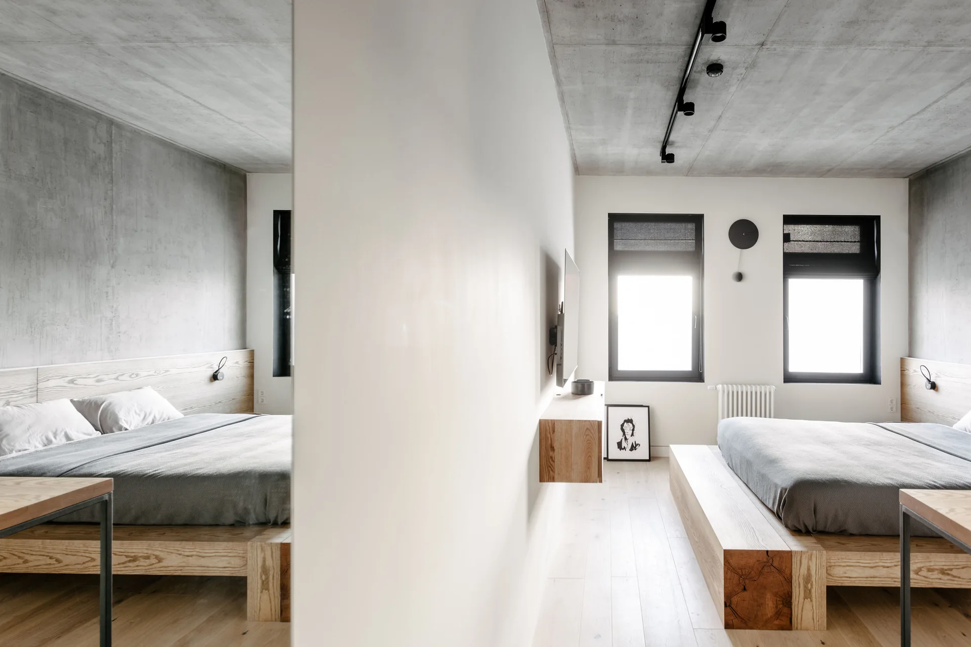 A minimalist bedroom features a chunky wood platform bed, a raw concrete ceiling with black track lights, and a floor-to-ceiling mirror.