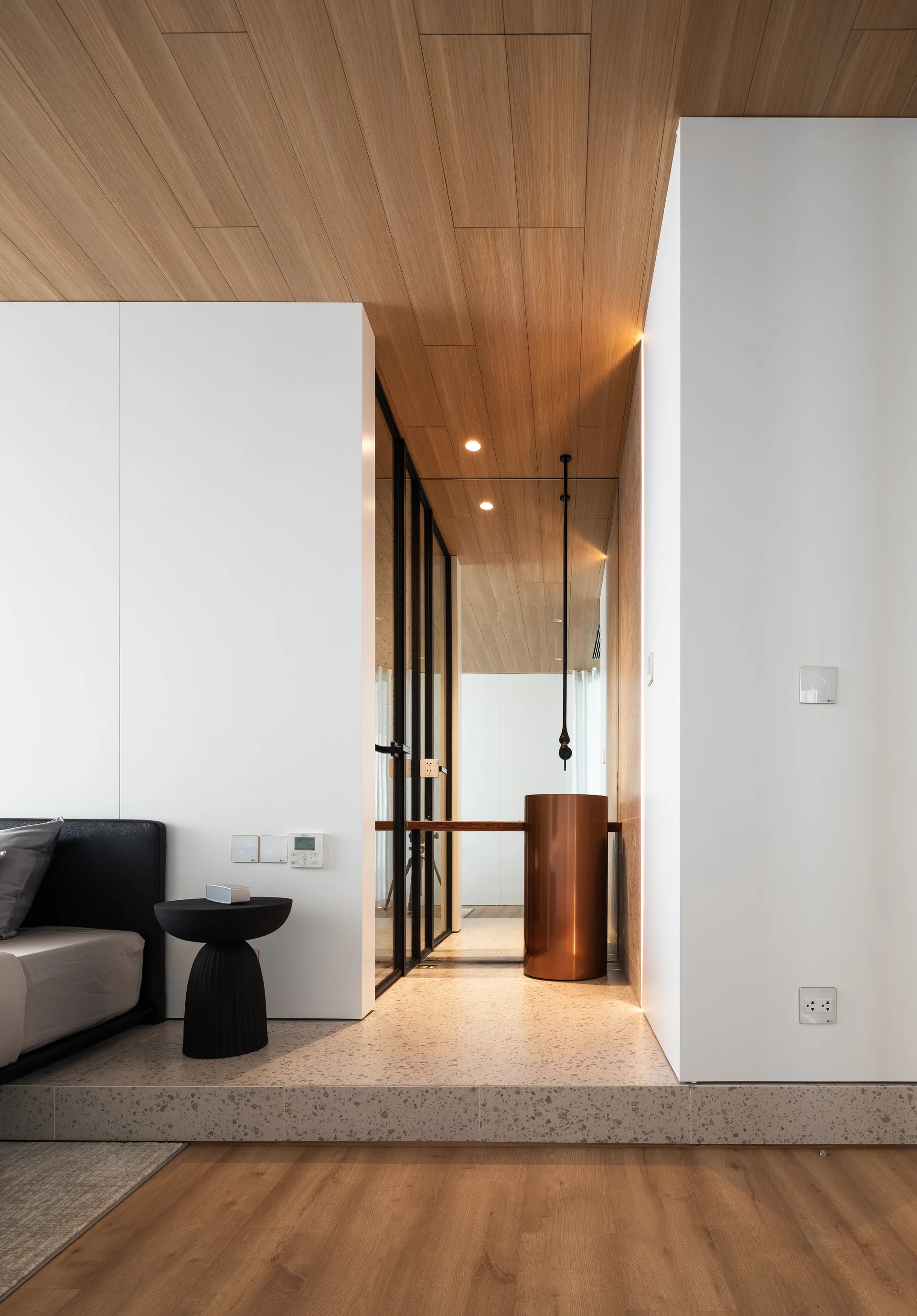 A modern bedroom features a raised terrazzo platform leading to a black-framed glass wall and a cylindrical copper pedestal sink.