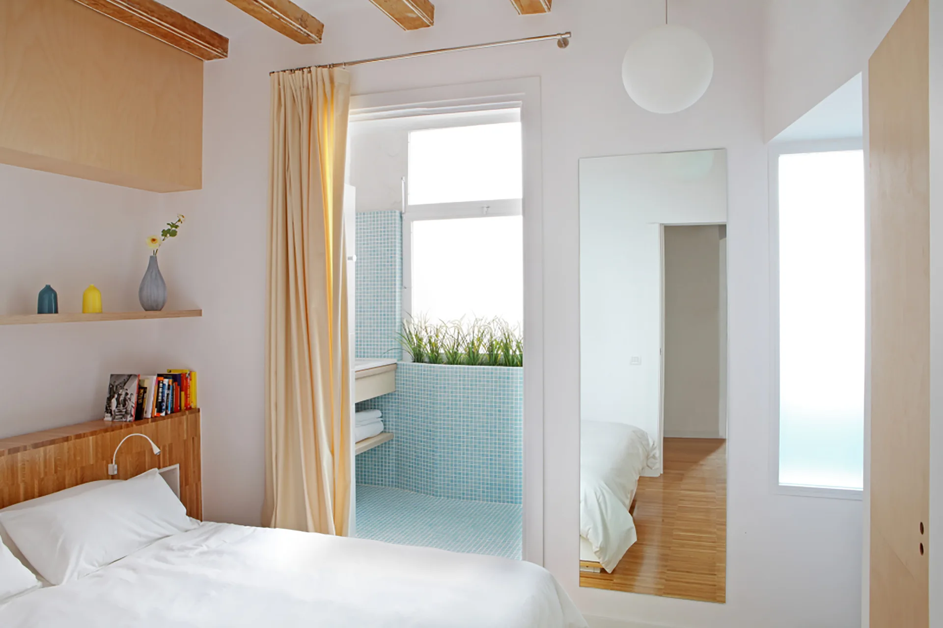 A minimalist bedroom featuring a light wood headboard, white bedding, and a doorway opening to a blue mosaic-tiled bathroom.