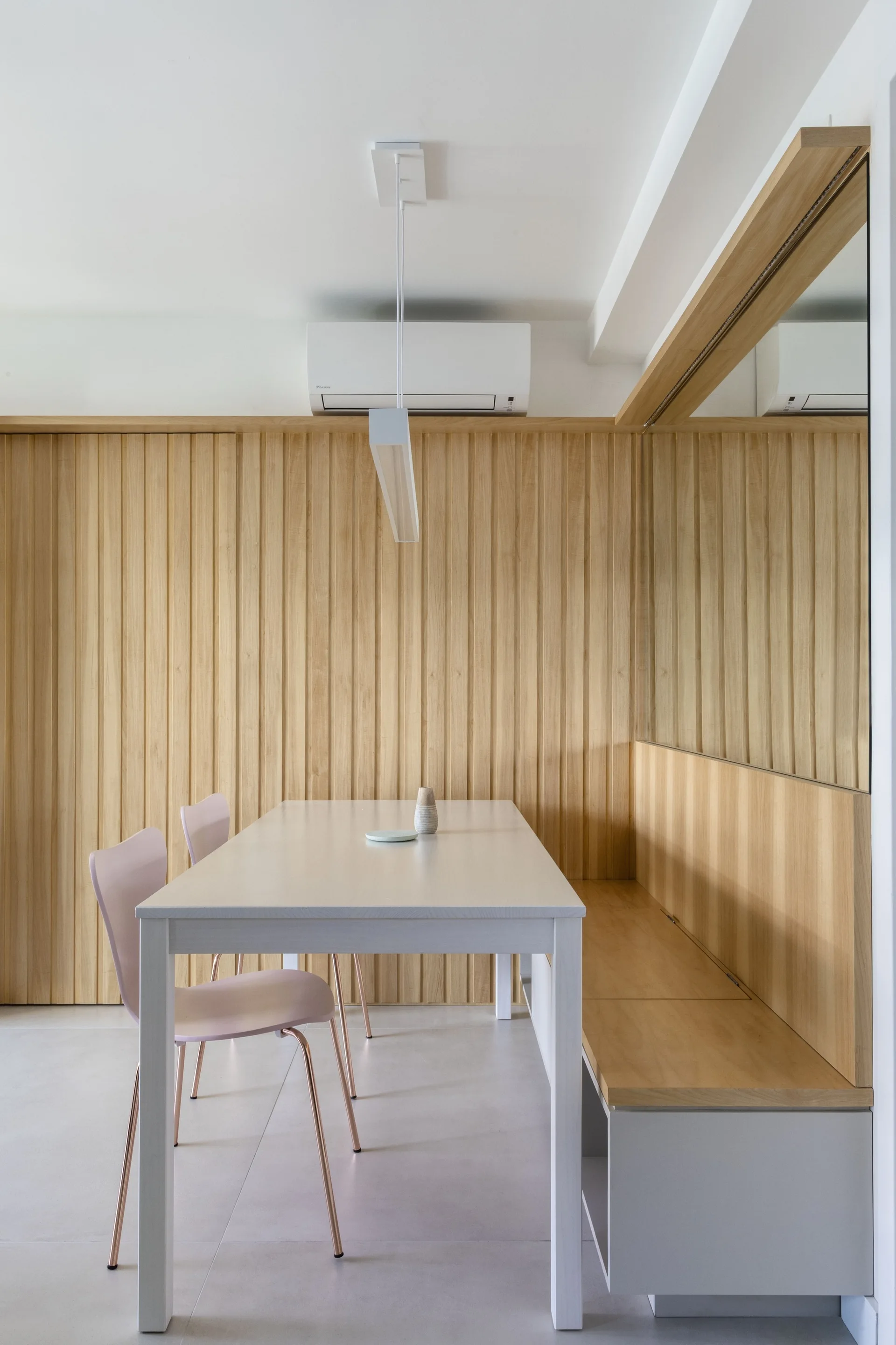 A minimalist dining room features a light wood table, pink chairs, and a built-in wooden bench against a vertical wood slat wall with a mirror.