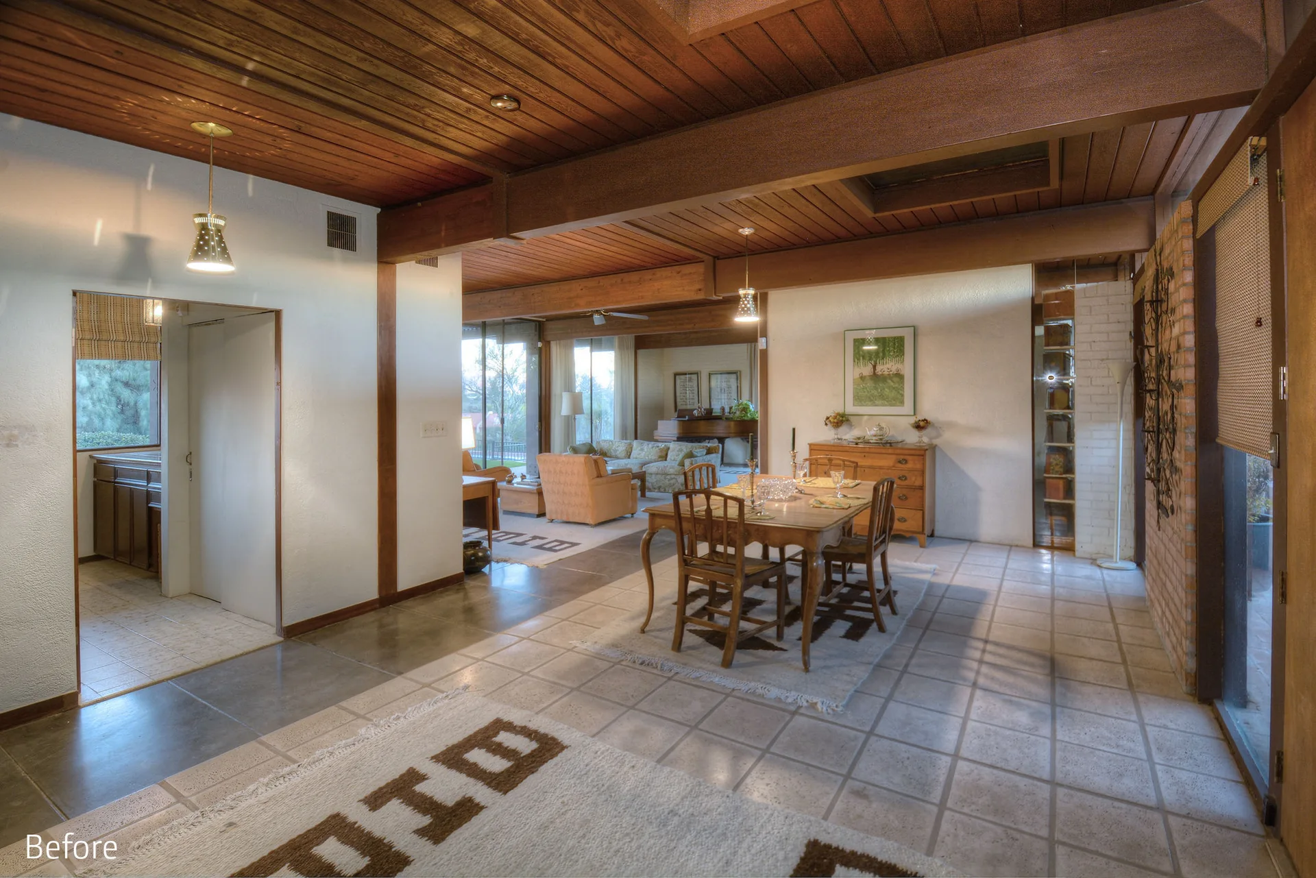 An open dining area features a wooden table and chairs on a patterned rug under a dark wood paneled ceiling with exposed beams.