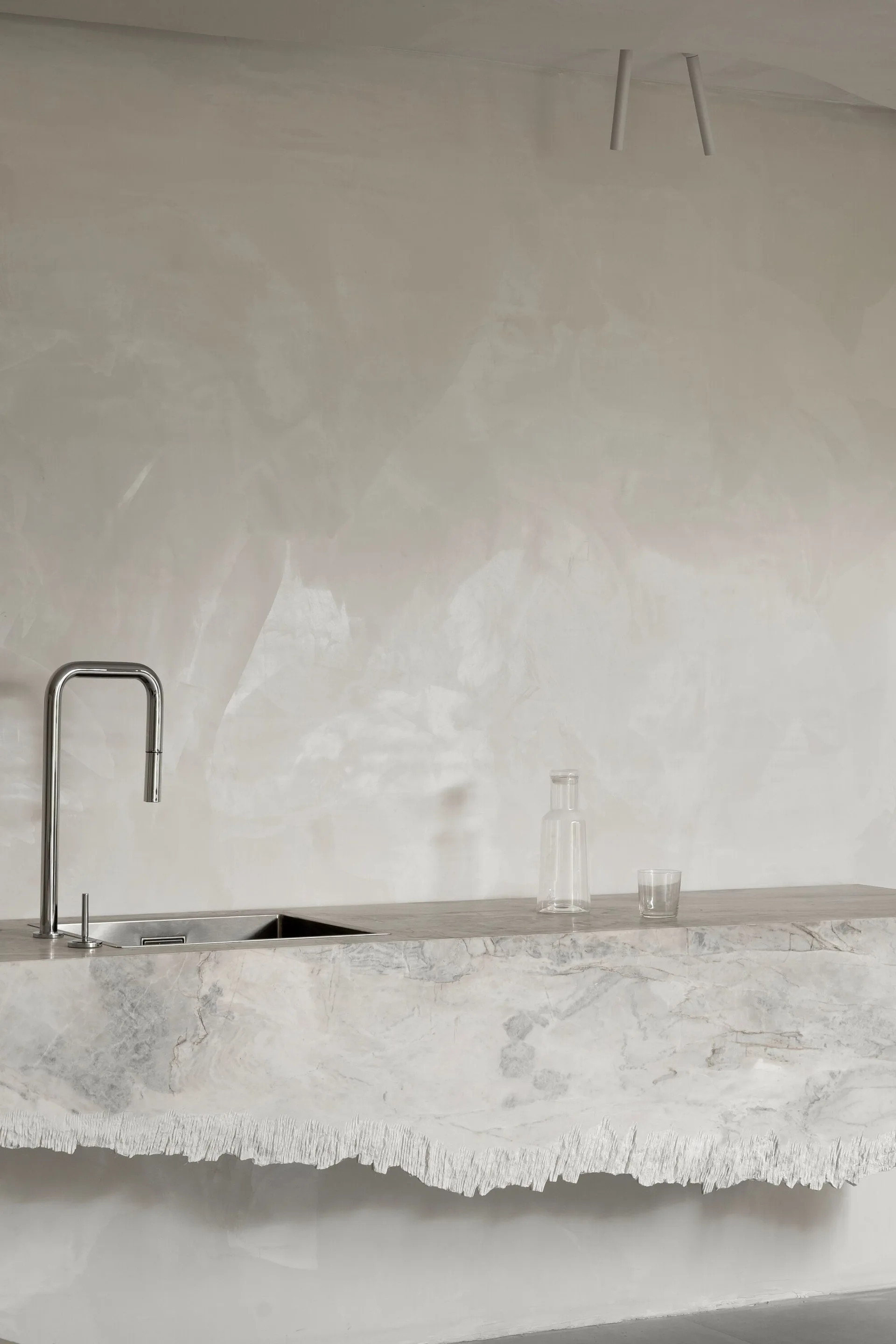 A modern kitchen sink set in a grey-veined marble countertop with a rough, unfinished lower edge against a matte off-white plaster wall.