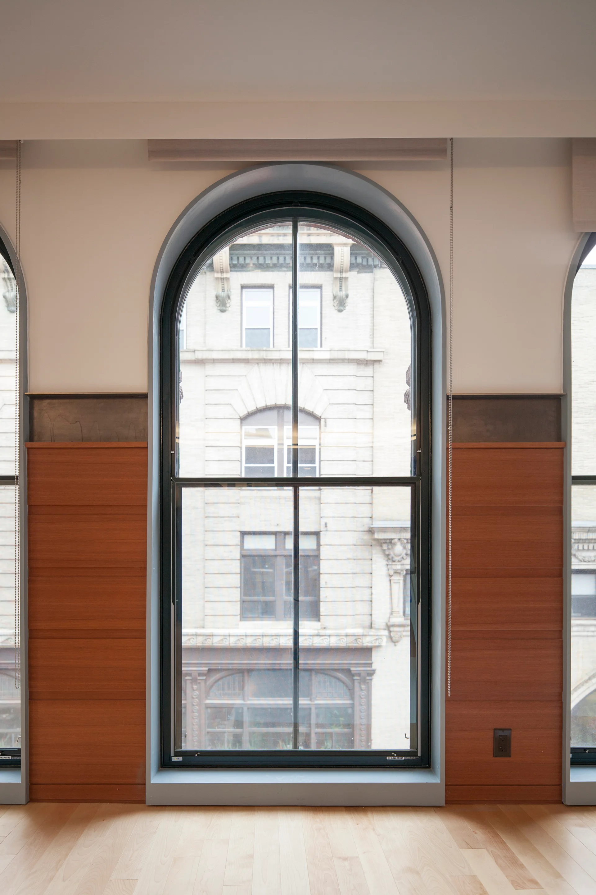 An interior view of a tall arched window framed by a dark gray trim, featuring horizontal wood wall paneling and a light wood floor.