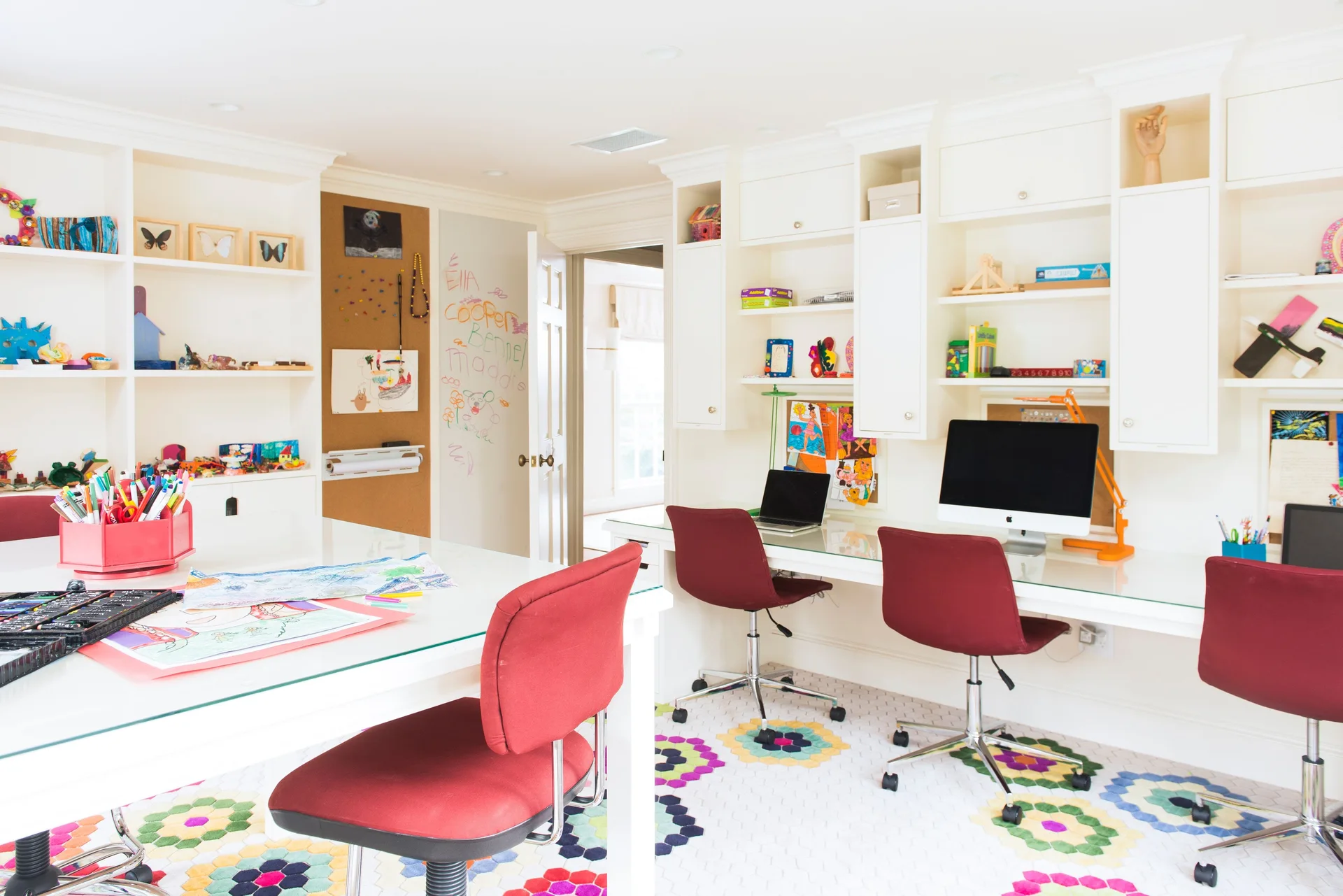 A bright home office features a long white built-in desk with red chairs, open shelves, and a colorful hexagonal patterned rug.