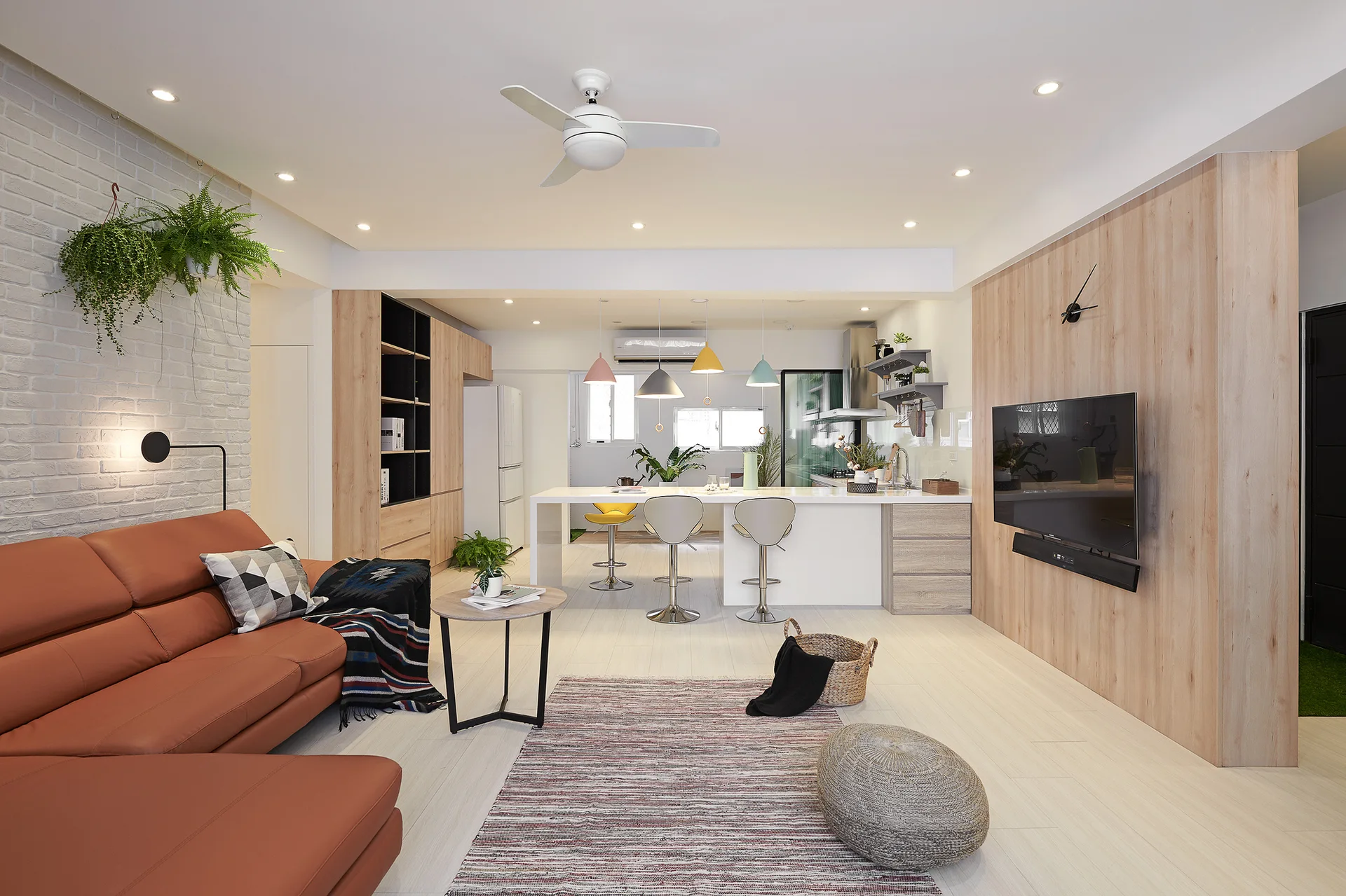 An open-plan living room features a brown leather sofa, a painted brick wall, and a wood-paneled accent wall facing a kitchen island.