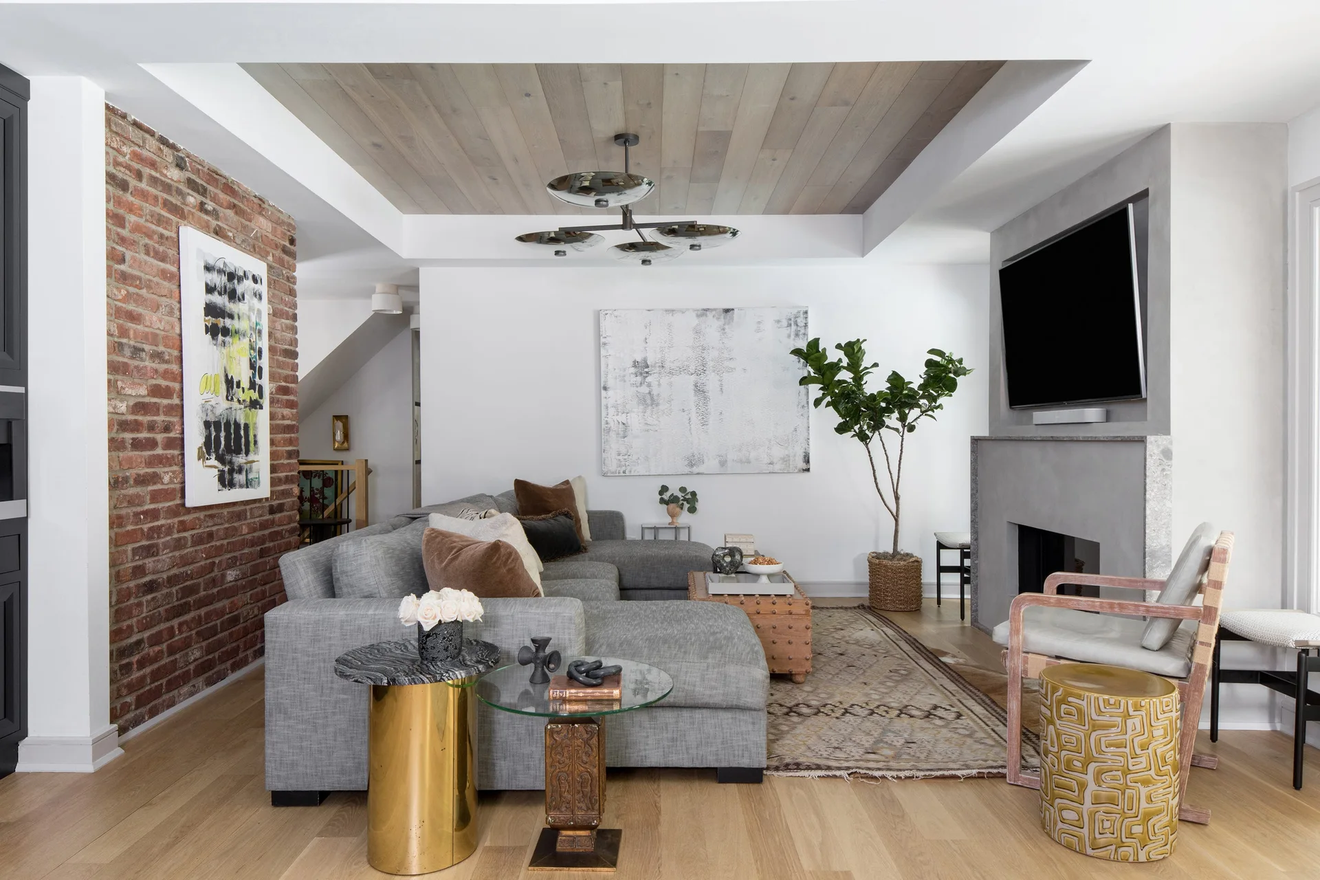 A modern living room featuring a grey sectional sofa, an exposed brick wall, a concrete fireplace, and a wood plank tray ceiling.