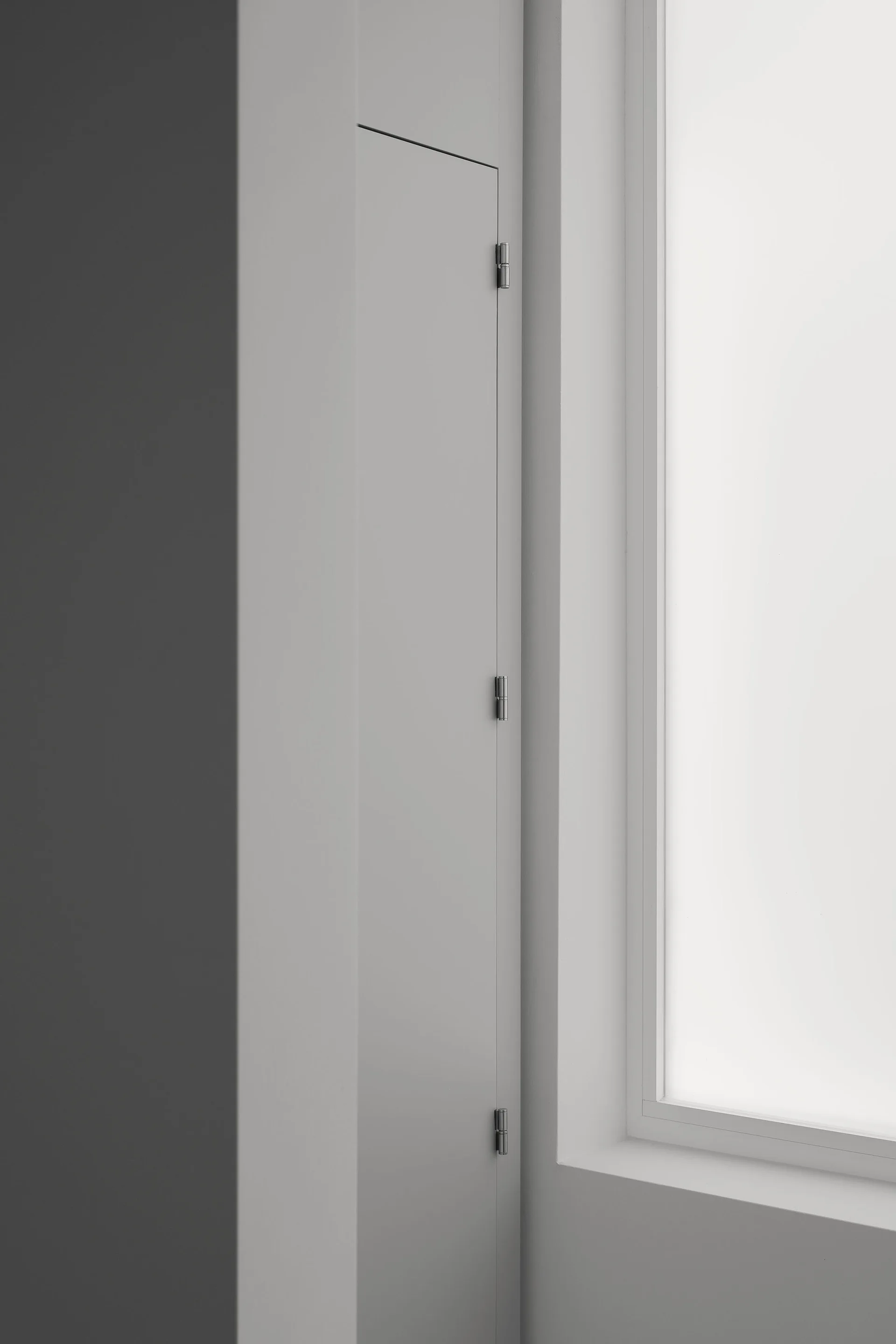 A close-up of a matte grey flush door panel with exposed silver metal hinges situated next to a bright window frame.