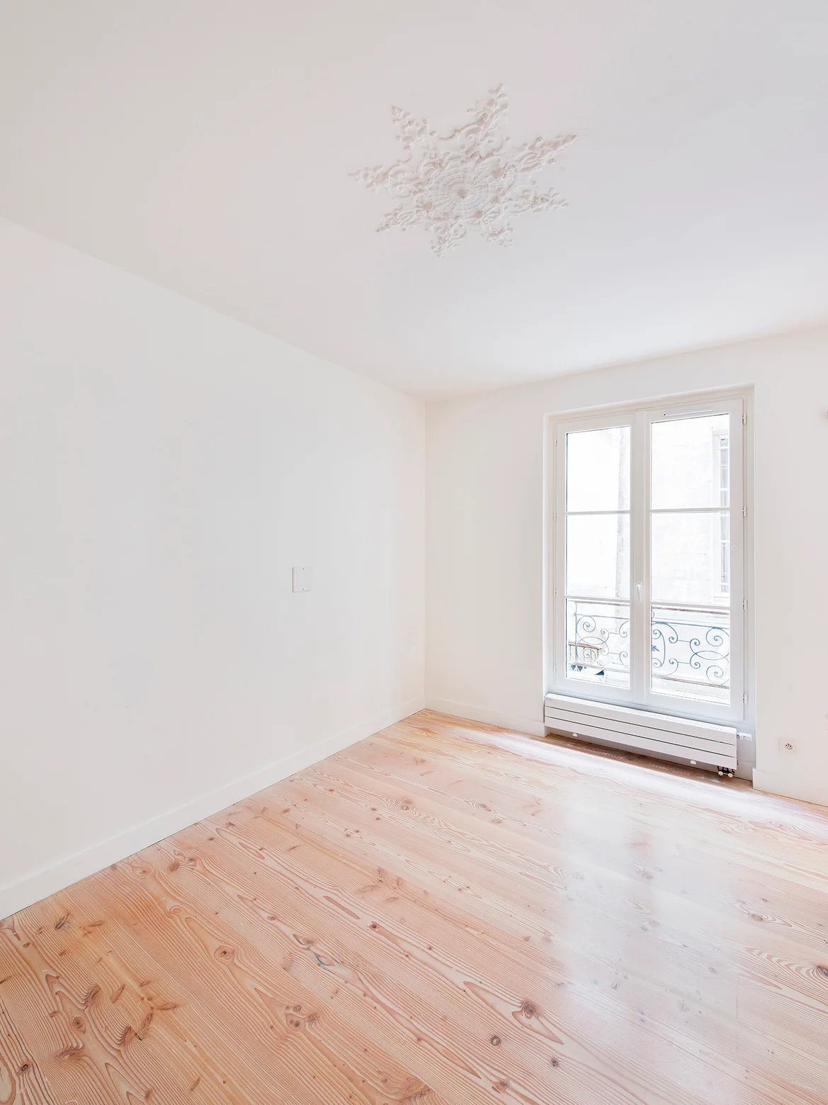 An empty room features light wood plank flooring, white walls, and an ornate plaster medallion on the ceiling next to a French window.