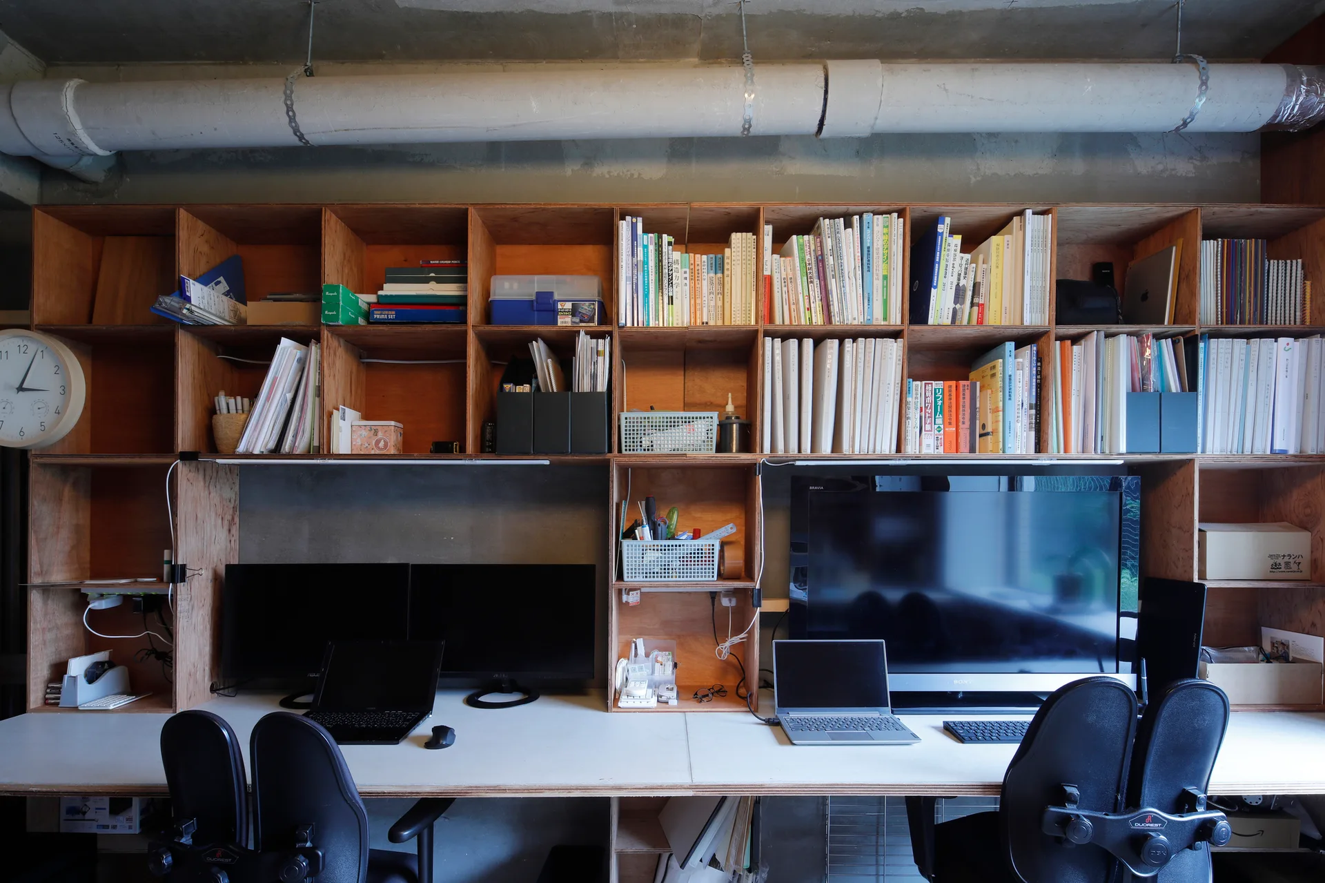 A home office workspace featuring a large plywood grid shelving unit mounted above a long white desk with two computer workstations.