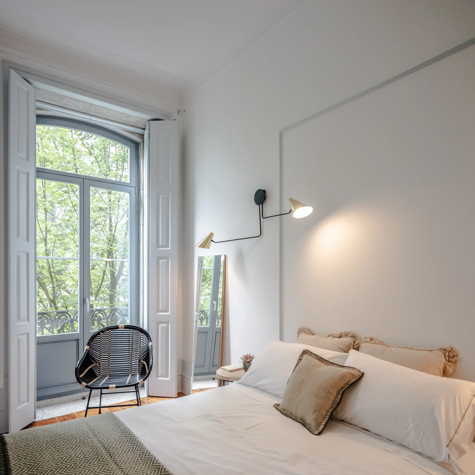 A bright bedroom featuring a bed with white linens, a black woven chair, and a double-armed wall sconce next to a large arched window with white shutters.