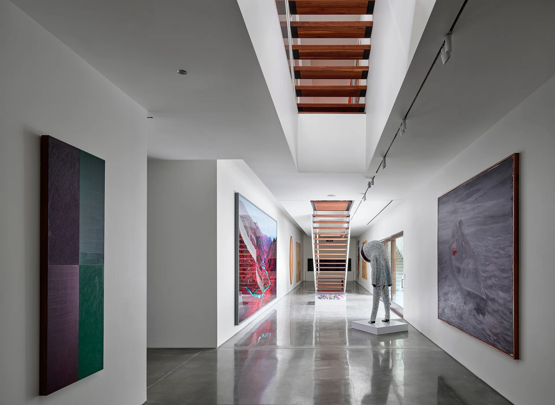 A minimalist white hallway features polished concrete floors, large modern art pieces, and a central floating staircase with wood treads.