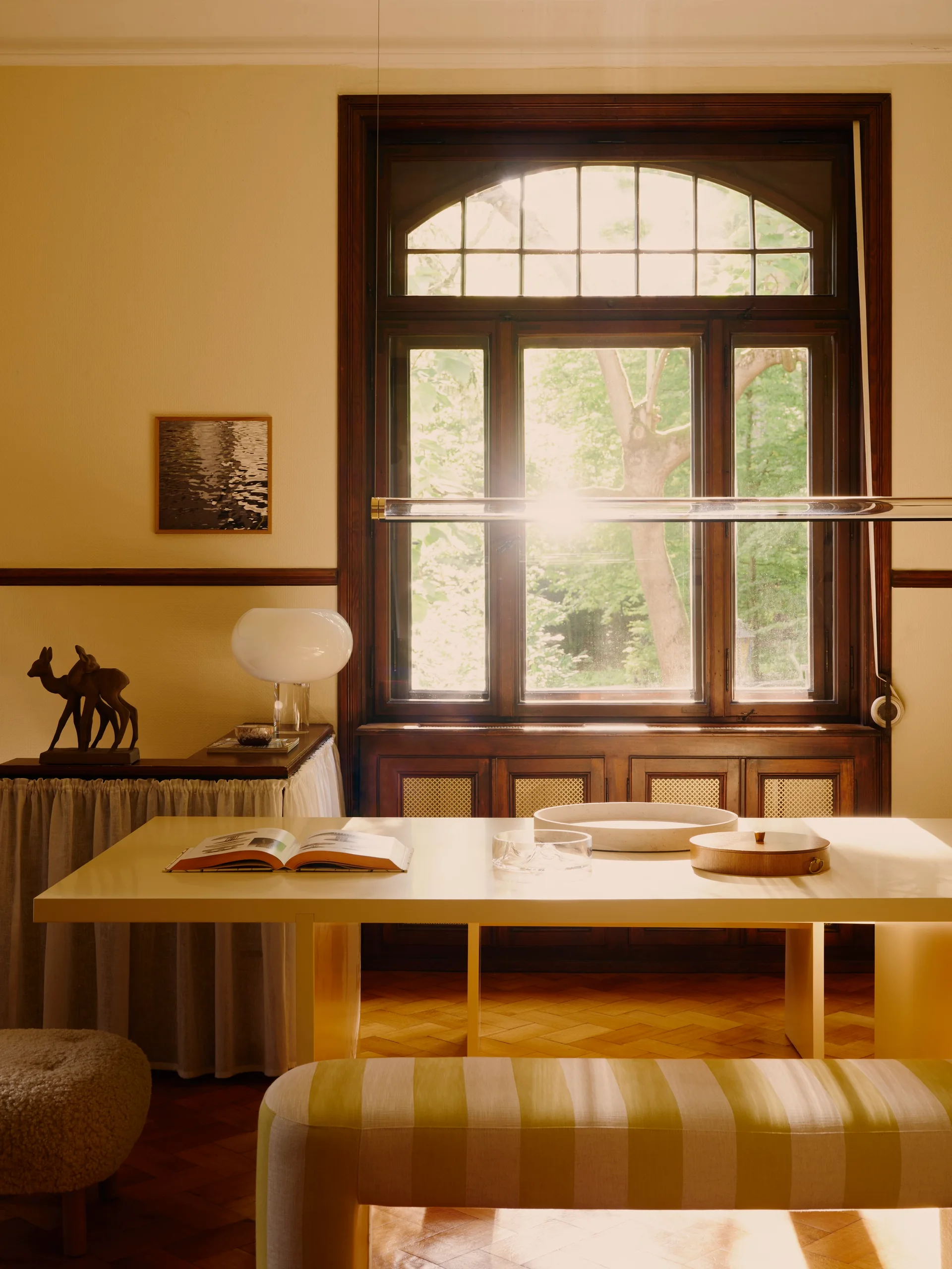 A sunlit dining room features a minimalist cream table, a striped upholstered bench, and a large dark wood arched window.