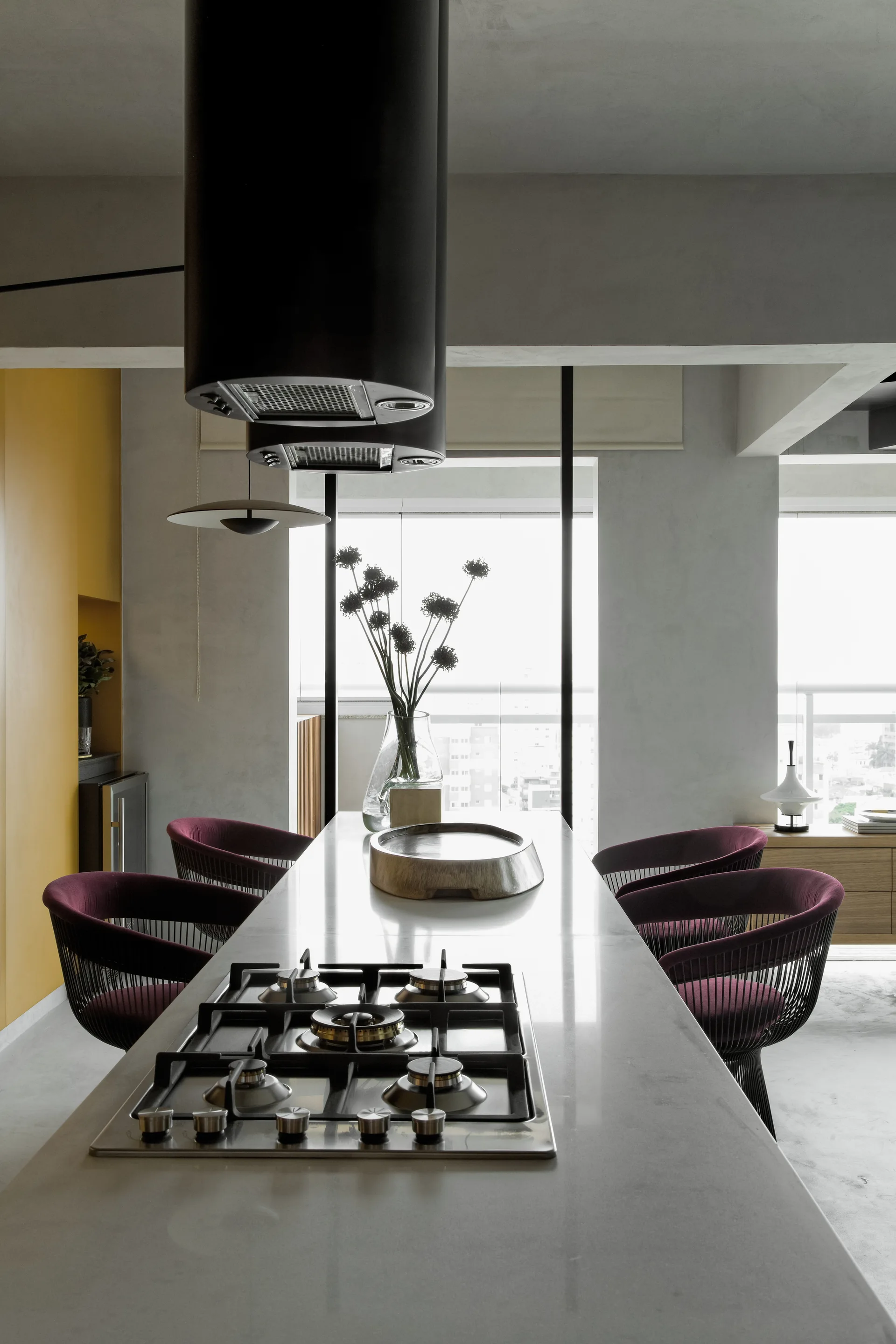A long concrete kitchen island with an integrated cooktop is surrounded by four deep purple chairs in front of a yellow accent wall.
